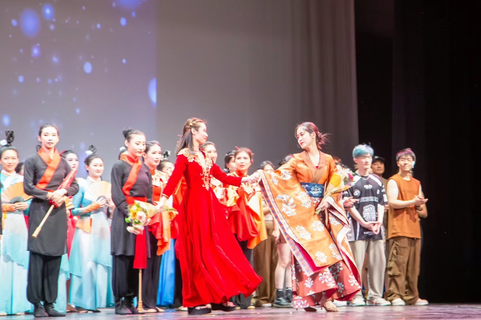 Group of children and adults performing on stage with colorful costumes and a starry background.