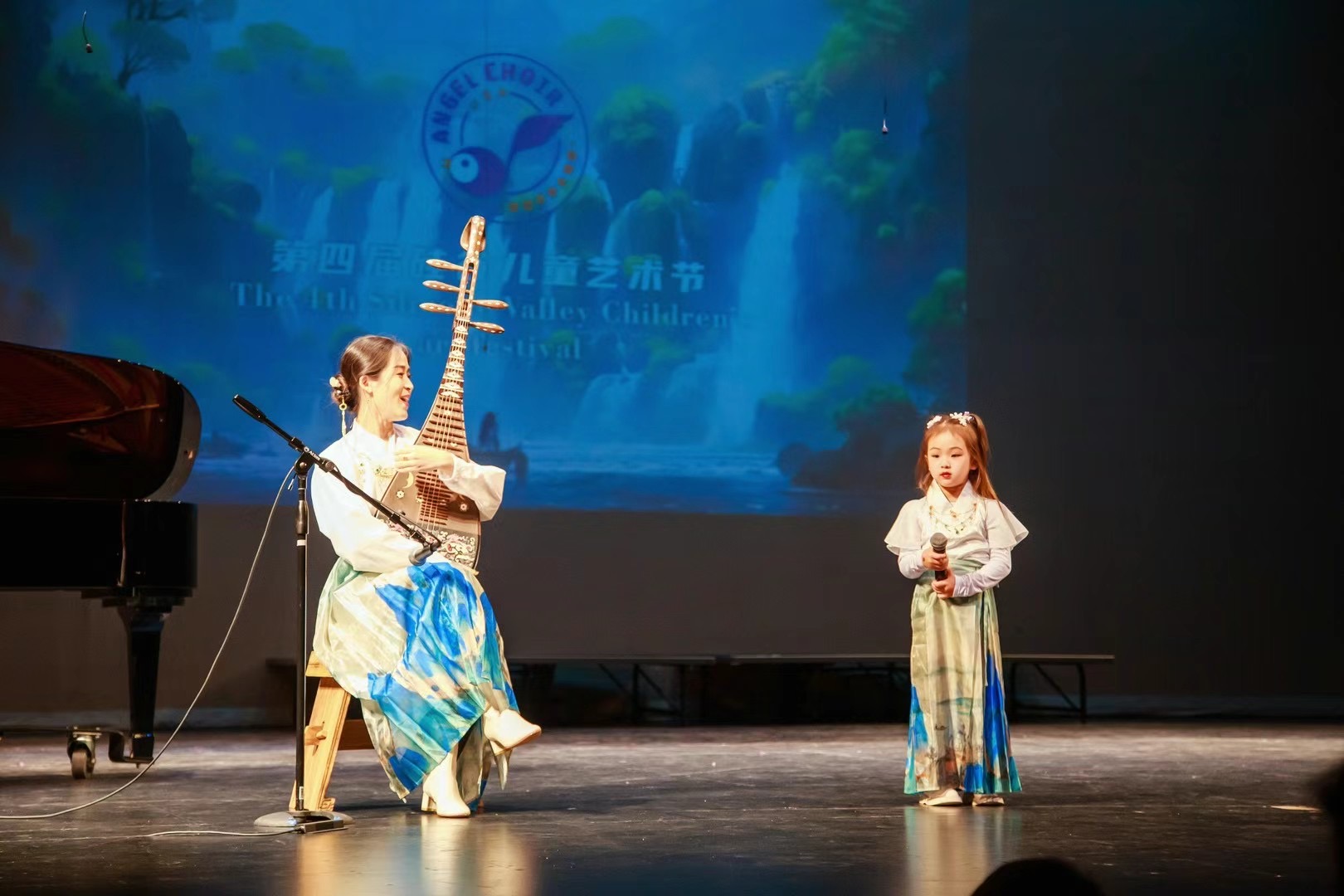 Two children performing on stage, one playing a traditional string instrument, the other holding a microphone, with a blue background screen.