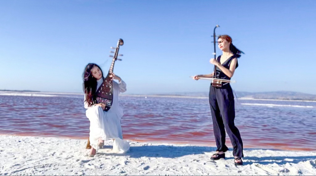 Two women playing musical instruments on a beach with ocean and sky in background.