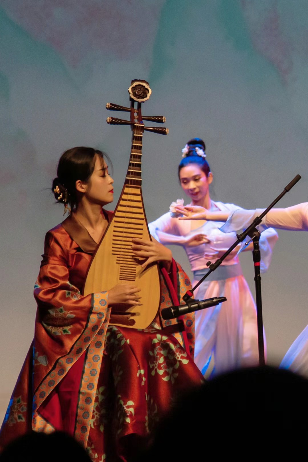 Two women perform on stage, one playing a traditional string instrument, the other dancing in colorful attire.