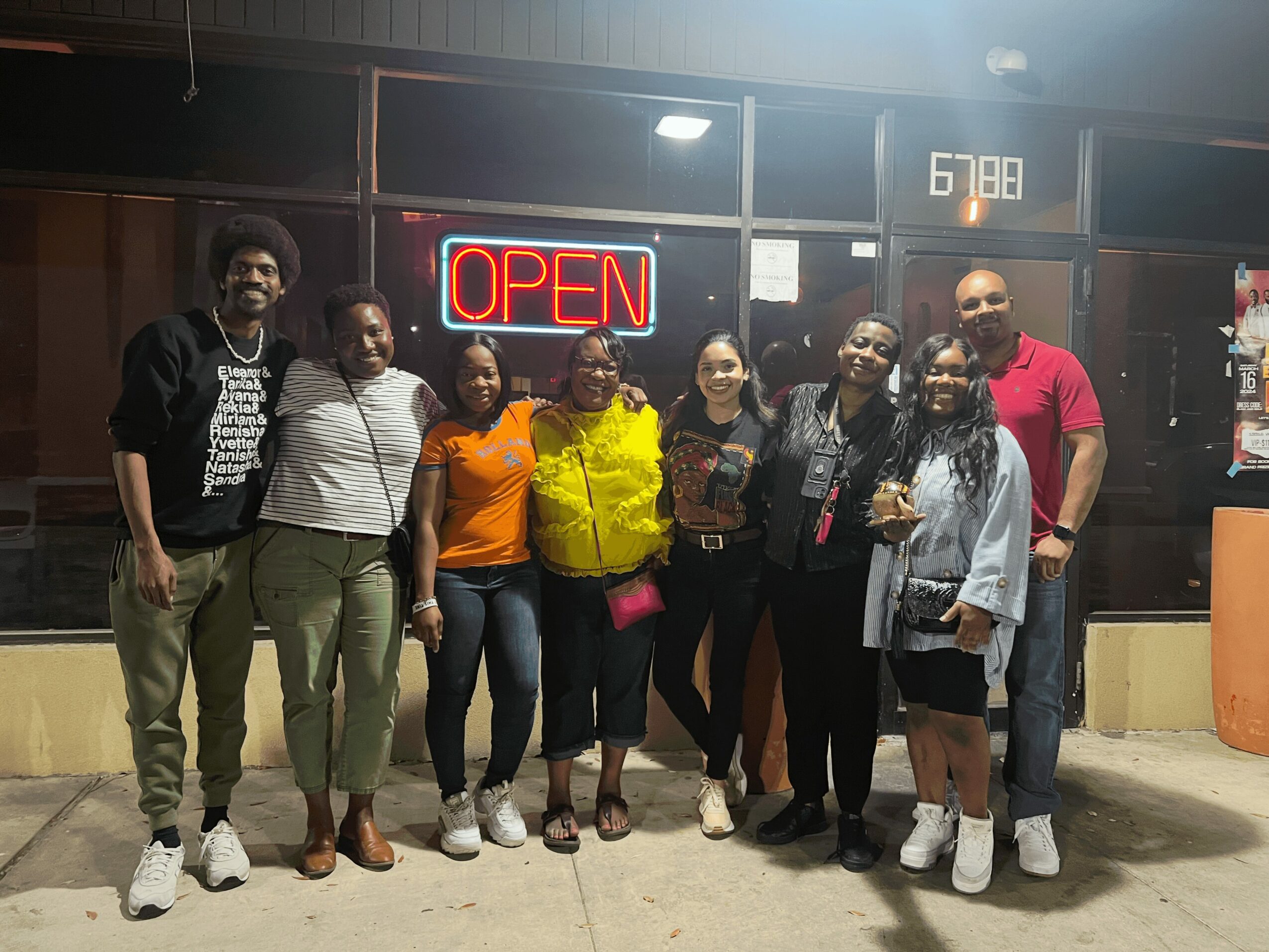 Group of nine people standing outside at night, some holding drinks, with an illuminated 'OPEN' sign behind them.