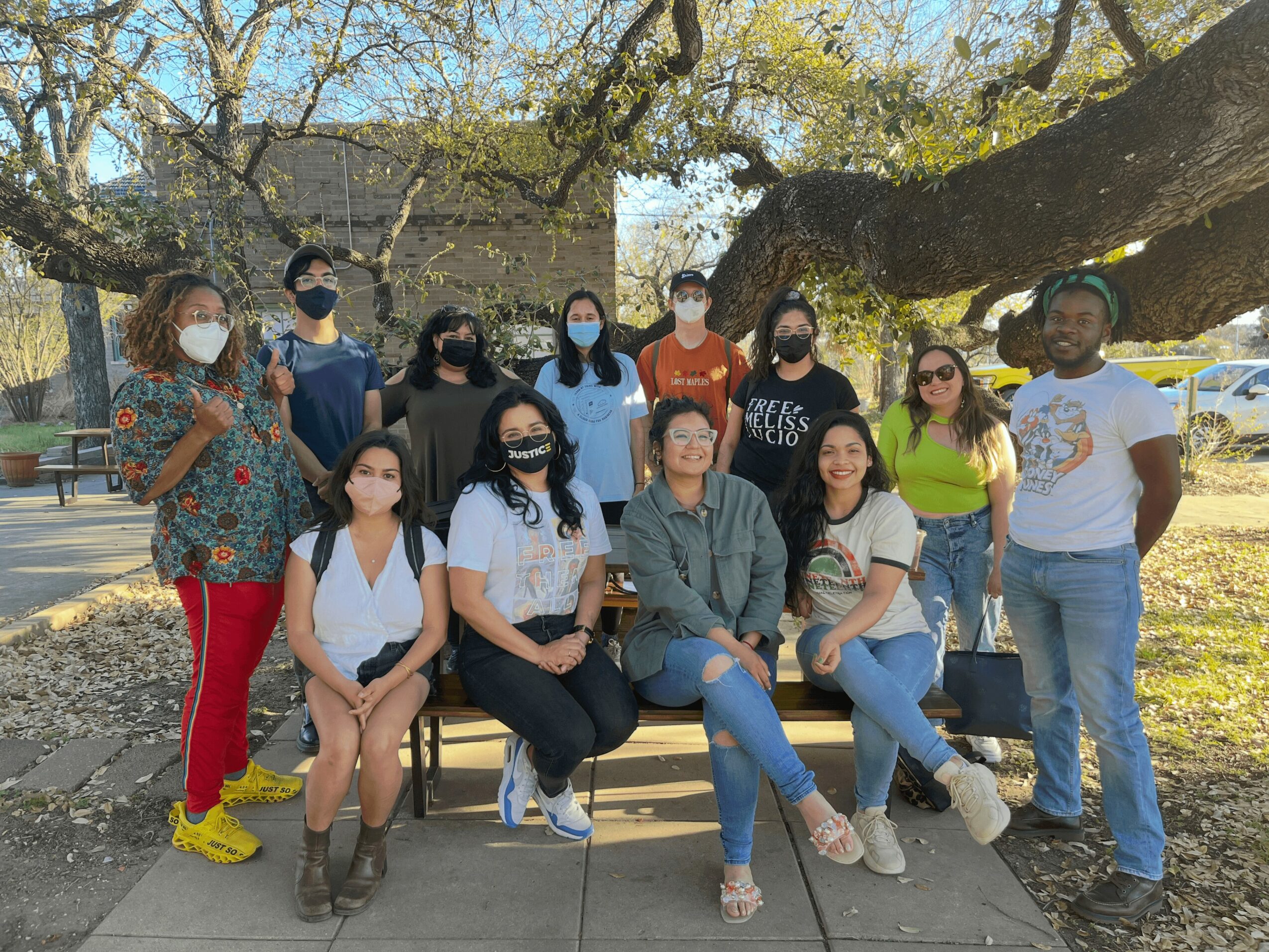 Group of people outdoors under a large tree, all wearing masks, posing for a photo.
