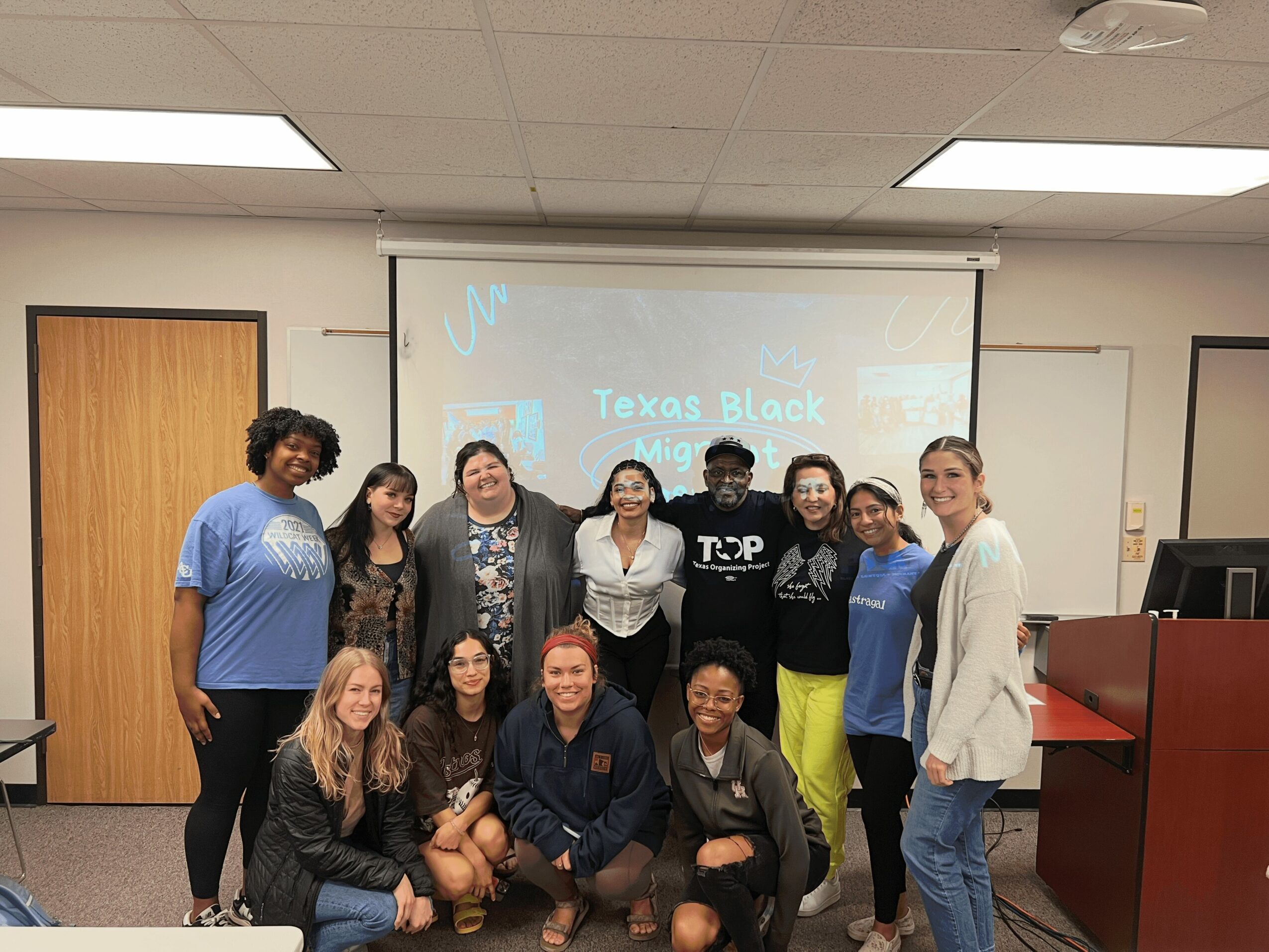 Group of ten people standing in a classroom, some smiling, with a projector screen behind them displaying text.