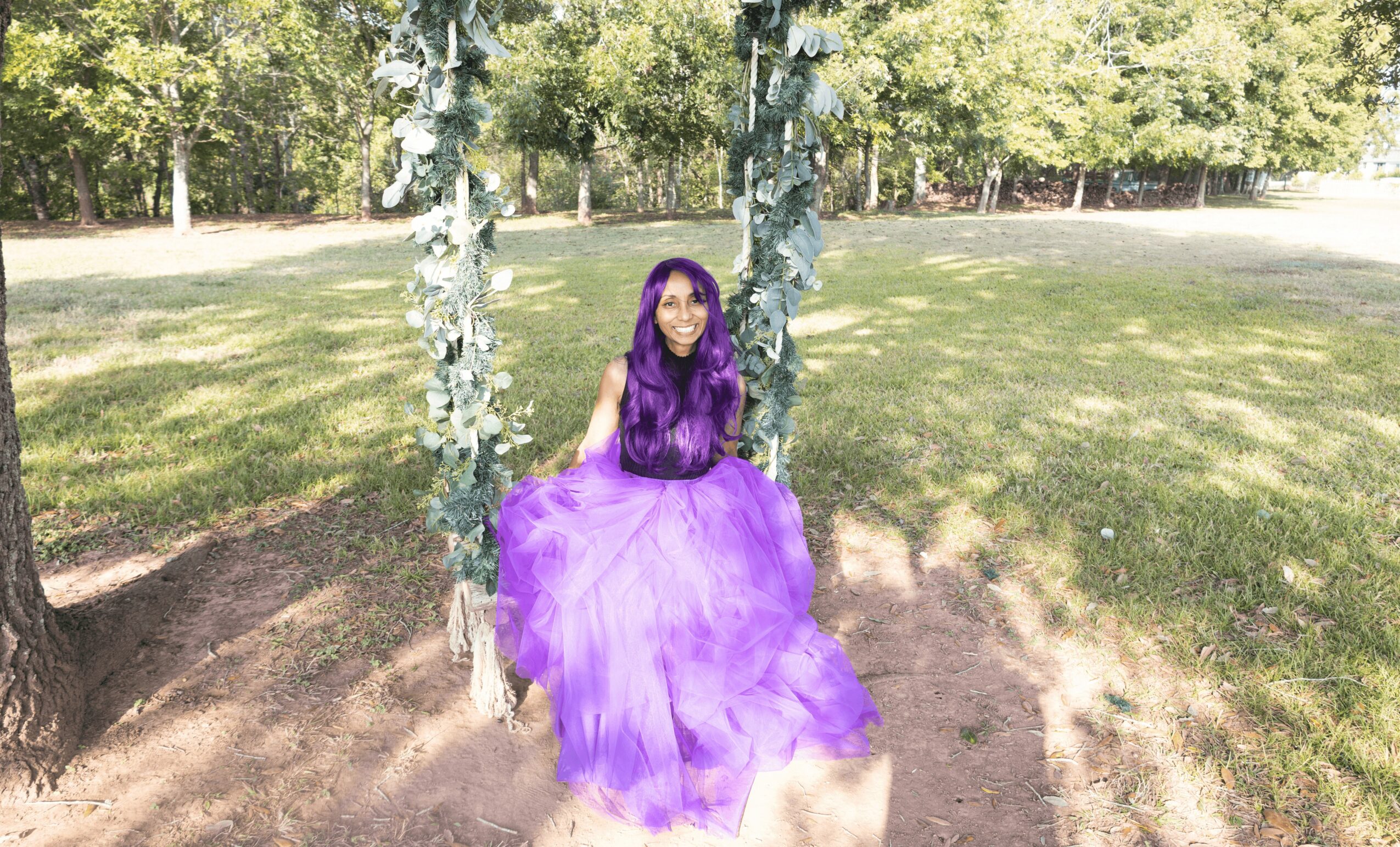 Girl in a purple dress sitting on the ground between two trees in a park.