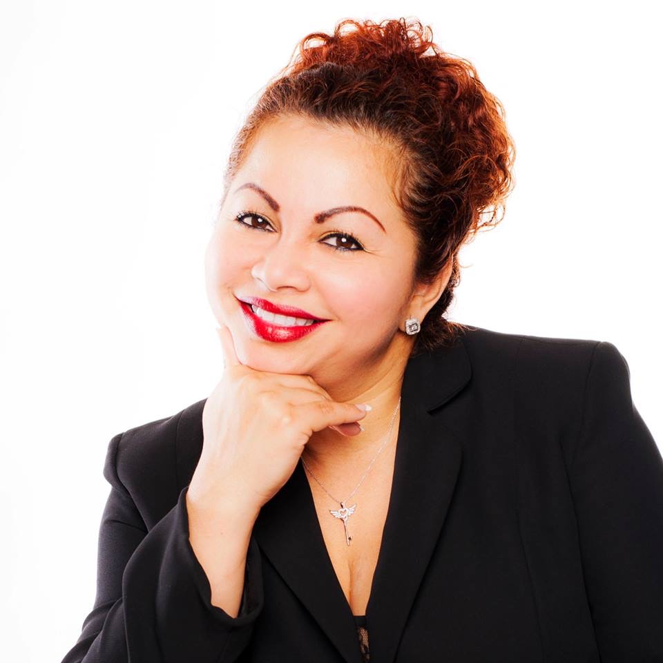 Woman with curly hair, smiling, wearing a black blazer and jewelry, resting her chin on her hand.