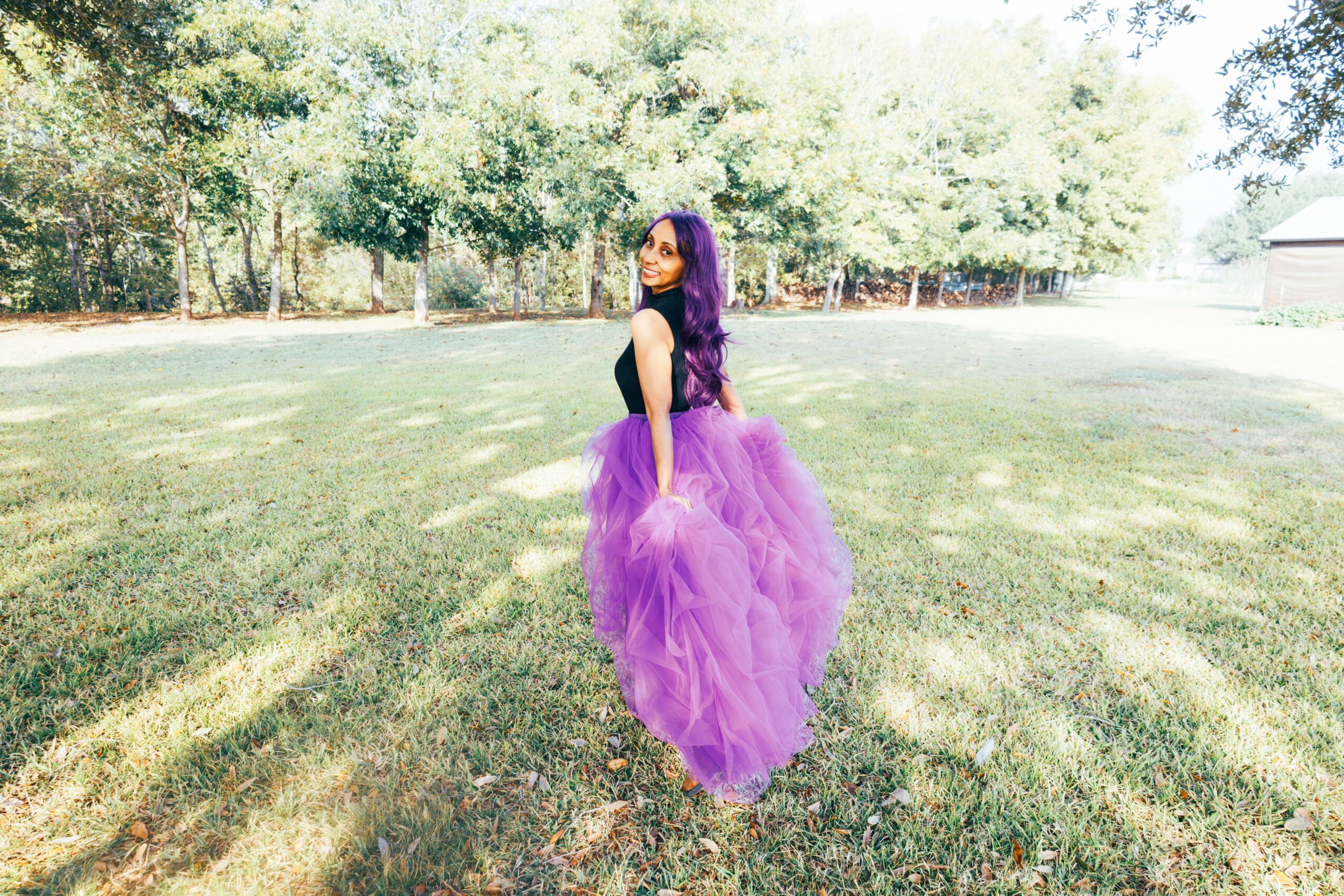 Girl in purple dress standing on grass with trees in background, facing sideways.