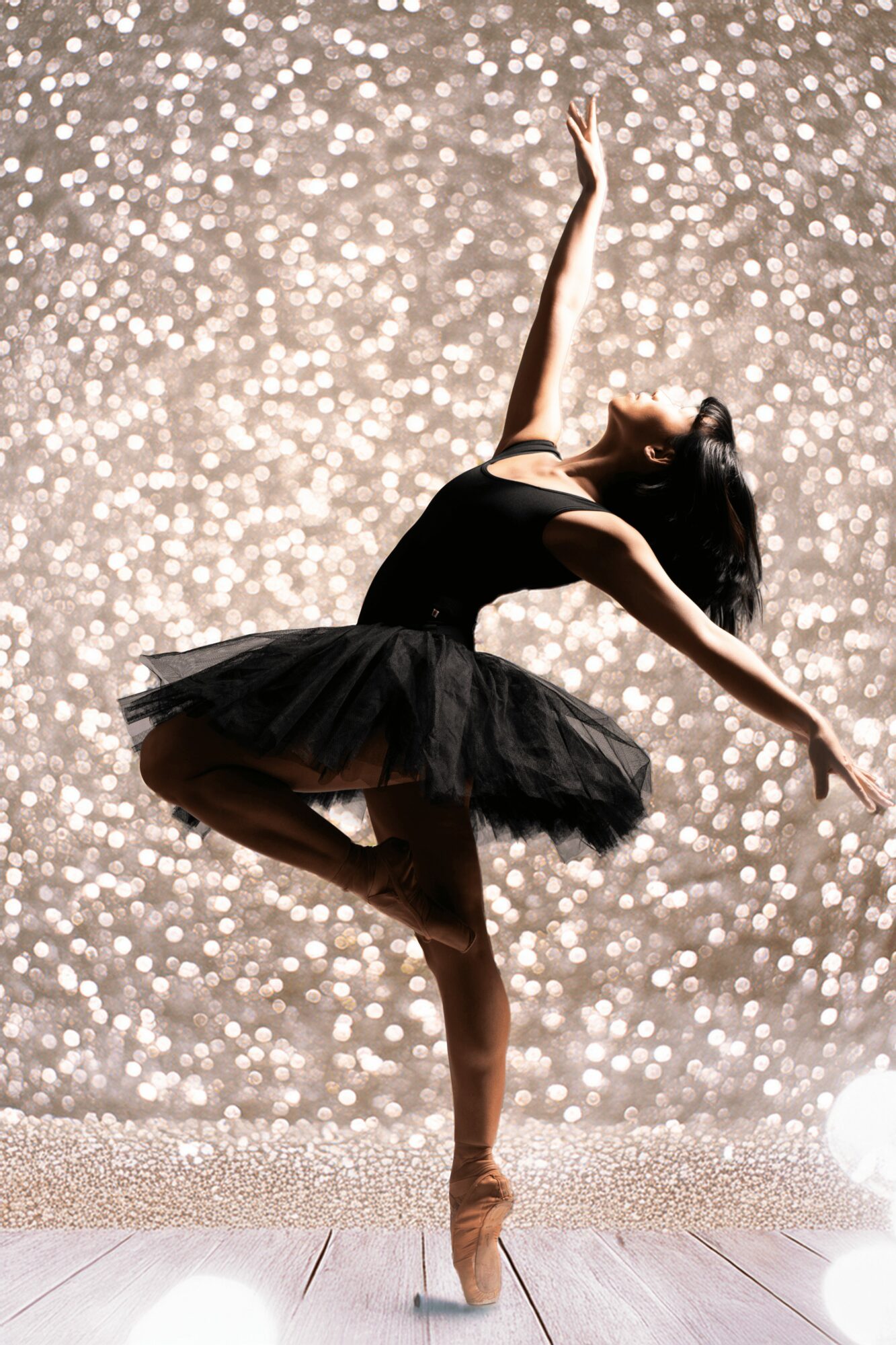 Ballet dancer in black tutu poses on stage with a glittery background, one leg raised and arms extended gracefully.
