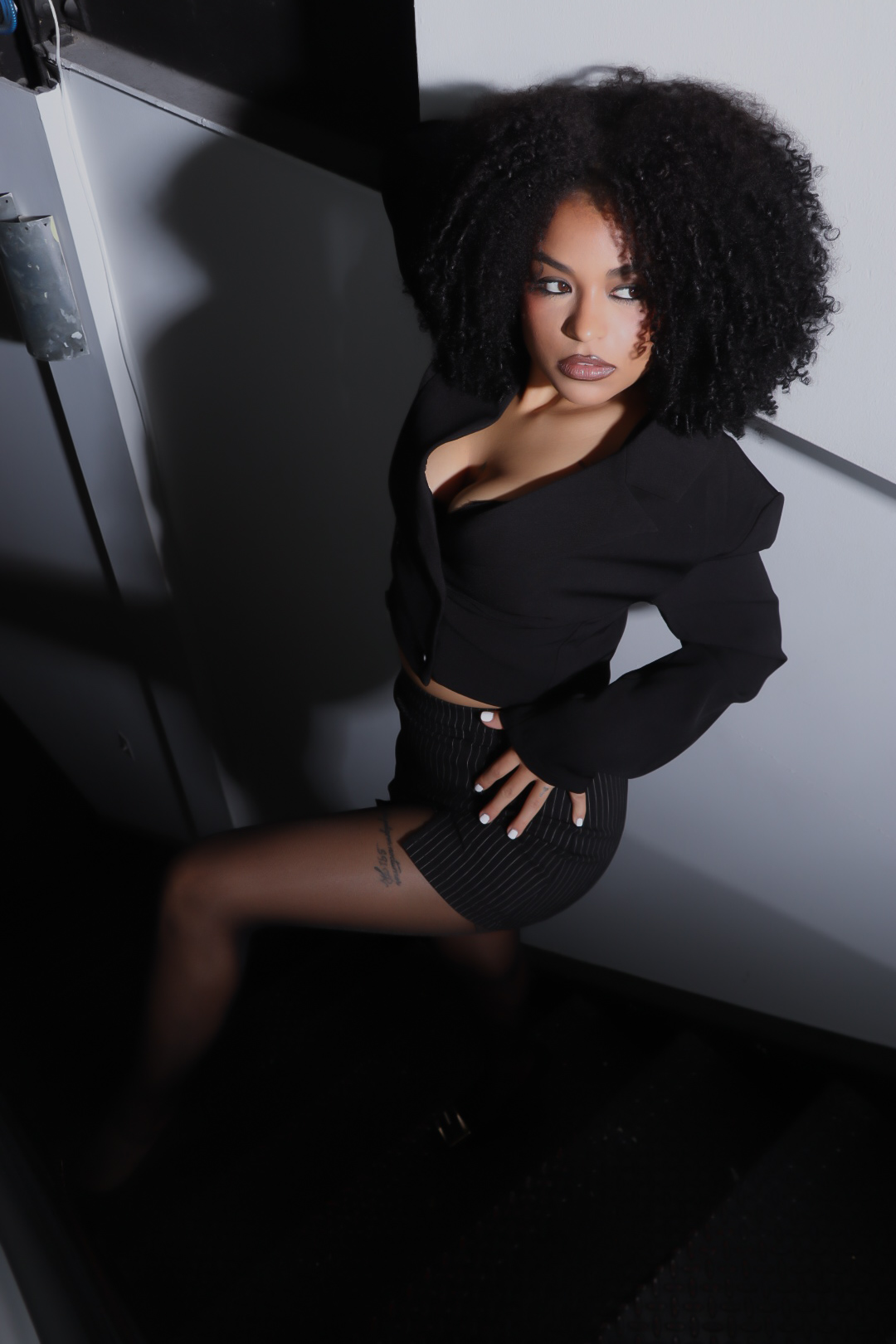 Woman with curly hair in black outfit sitting on a chair, looking at camera, in a room with shadows.