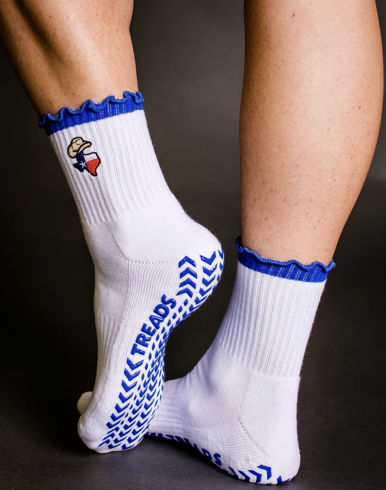 Two legs wearing white socks with blue ruffles and a logo, one sock slightly lifted, against a dark background.