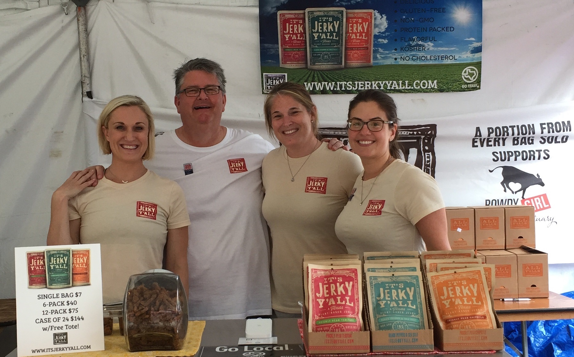 Four people standing behind a table with jerky products, smiling, in a tent at an event.