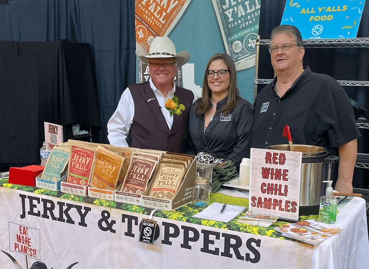 Three people behind a booth with jerky and toppings, smiling, at an indoor event.