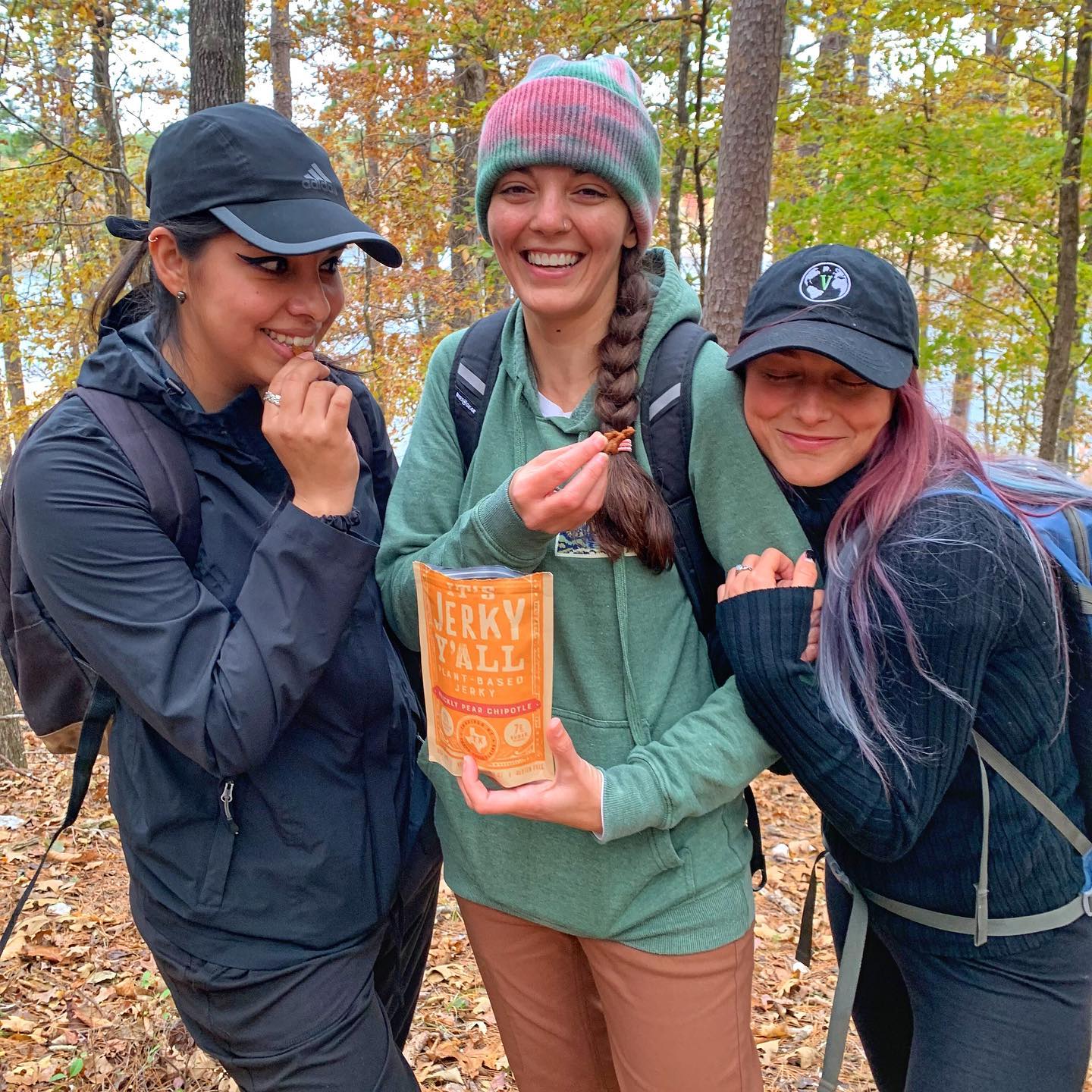 Three women outdoors in a forest, smiling, holding a bag of Jerky Fall snacks, wearing jackets and hats.