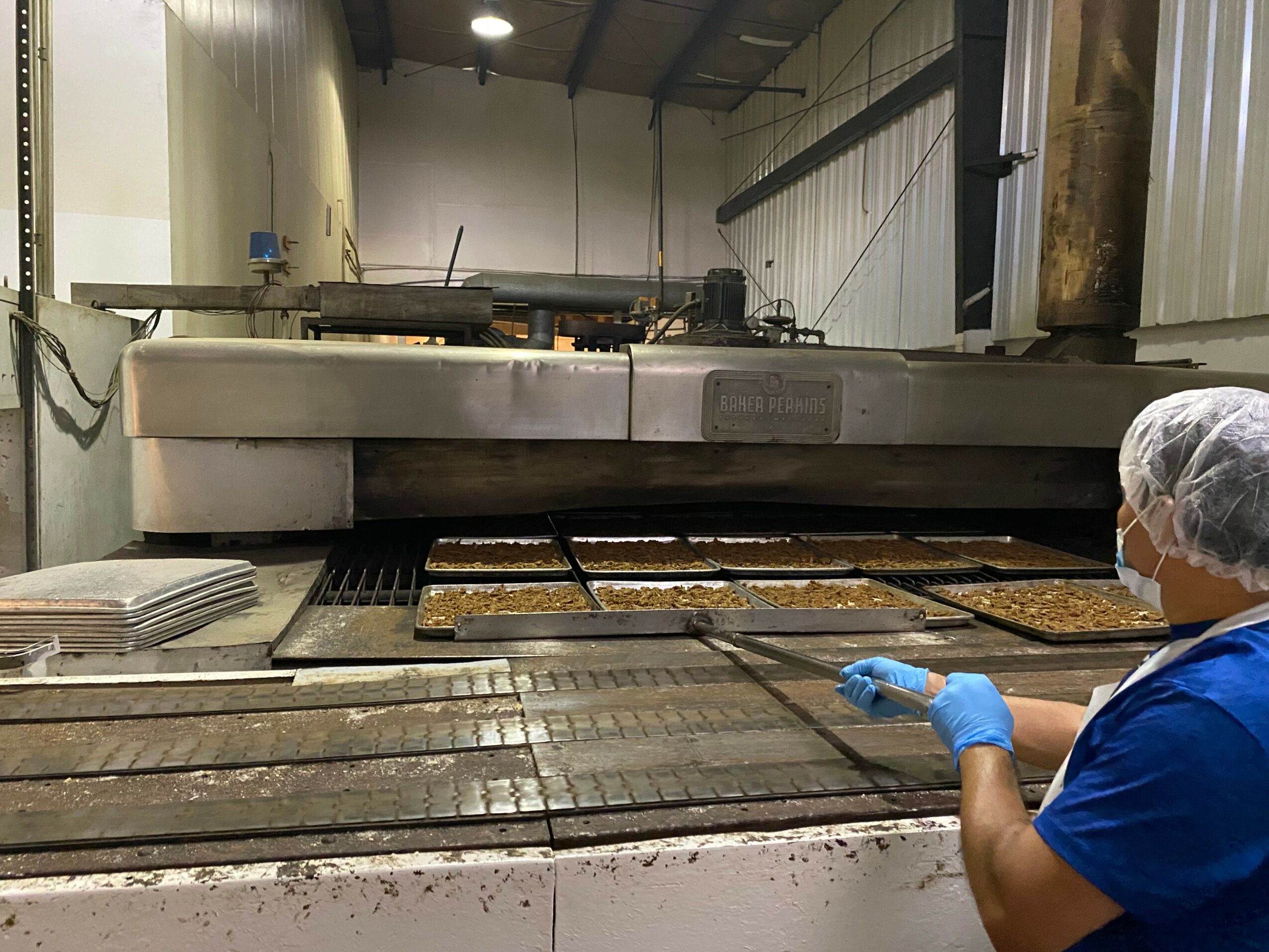 Worker wearing a hairnet and gloves inspects baked goods on a conveyor belt in a bakery.