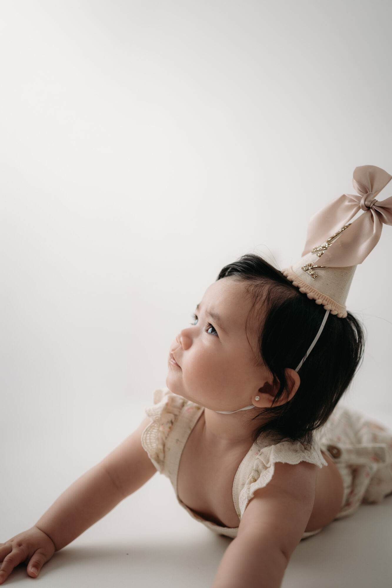 Baby lying on stomach wearing a party hat, looking upward, with a plain light background.