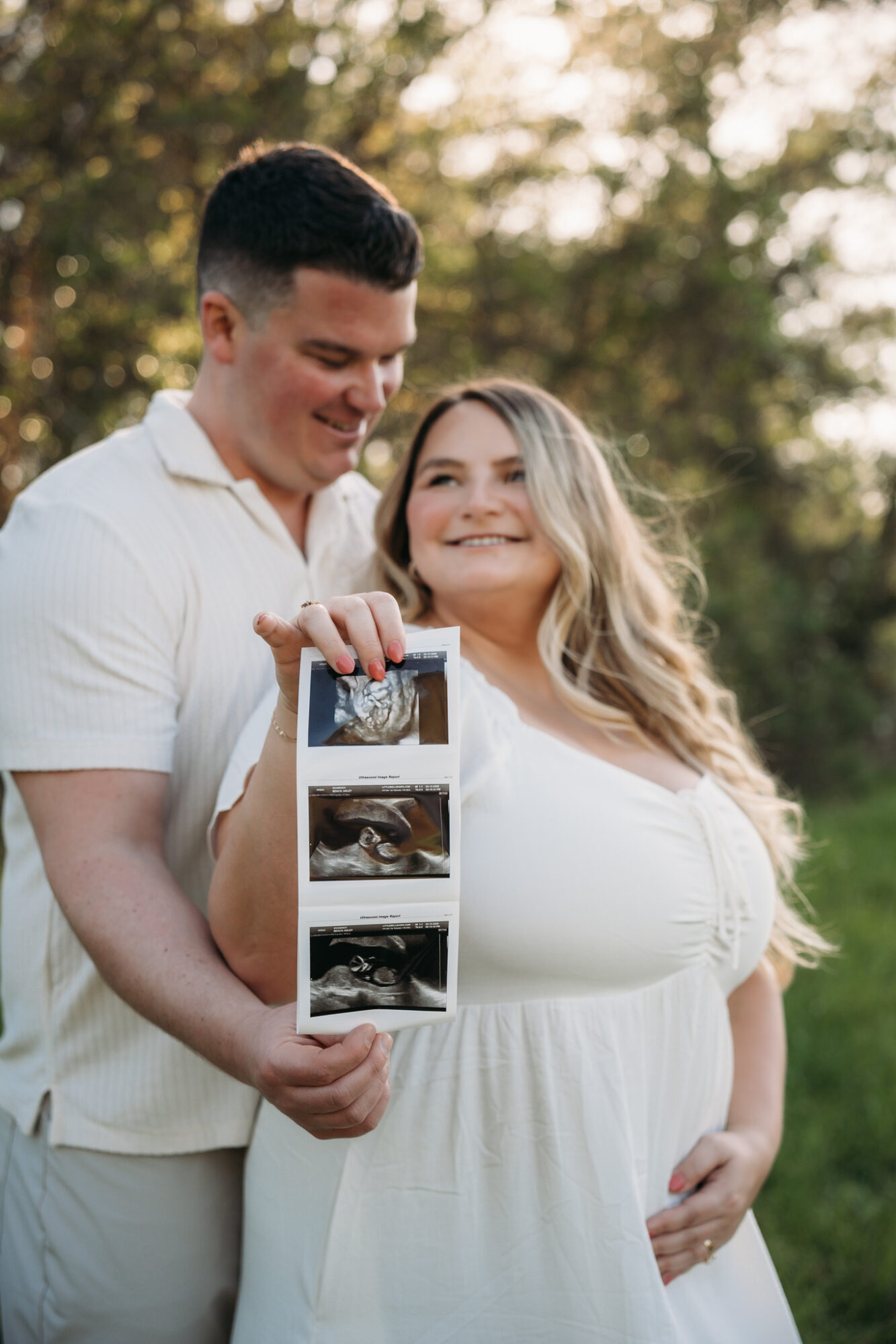 A couple outdoors holding ultrasound images, smiling, with trees in the background.