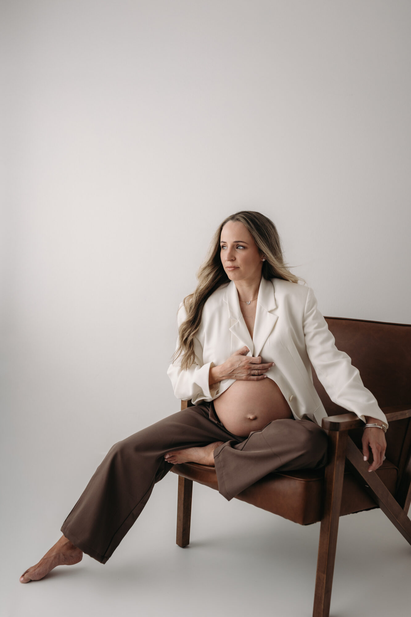 Pregnant woman sitting on a wooden chair, wearing a white blazer and brown pants, with a neutral background.