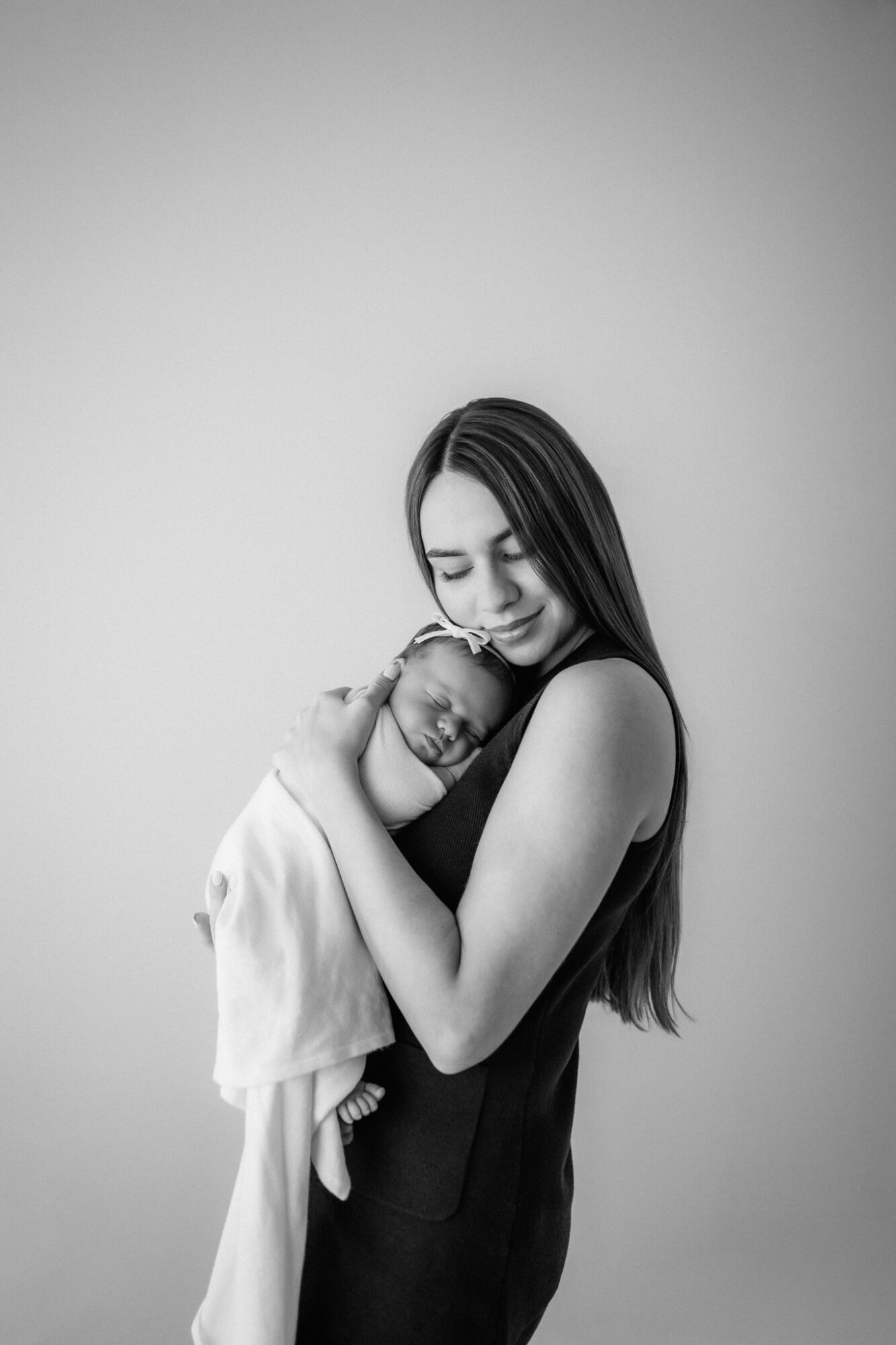 Woman holding a sleeping baby close, smiling gently, against plain background.