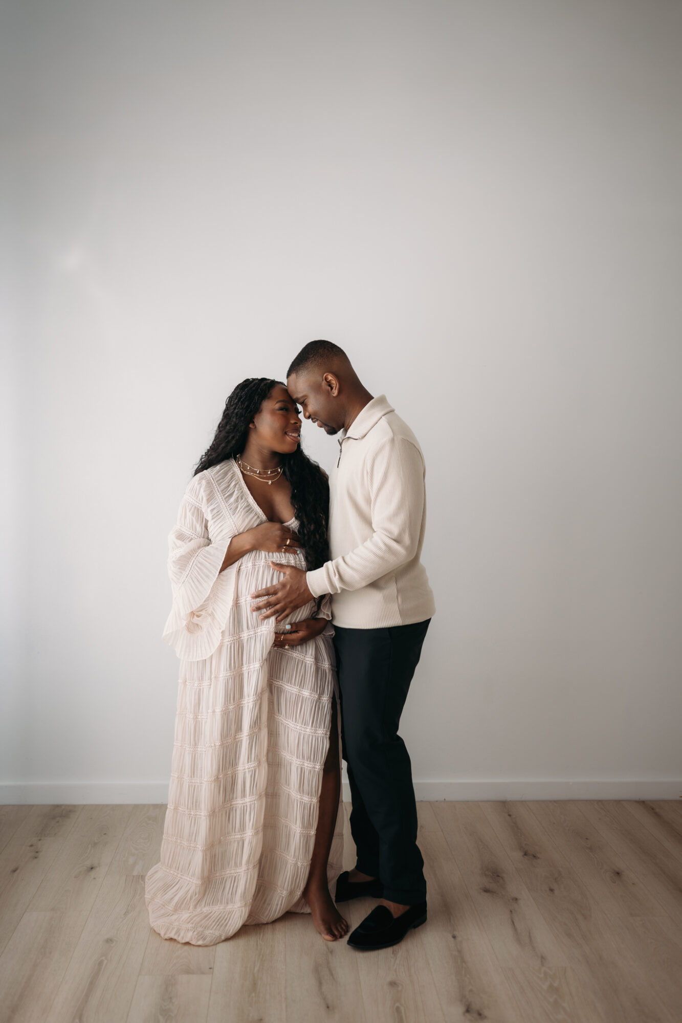 A couple stands close together, holding hands and looking at each other, against a plain light background.