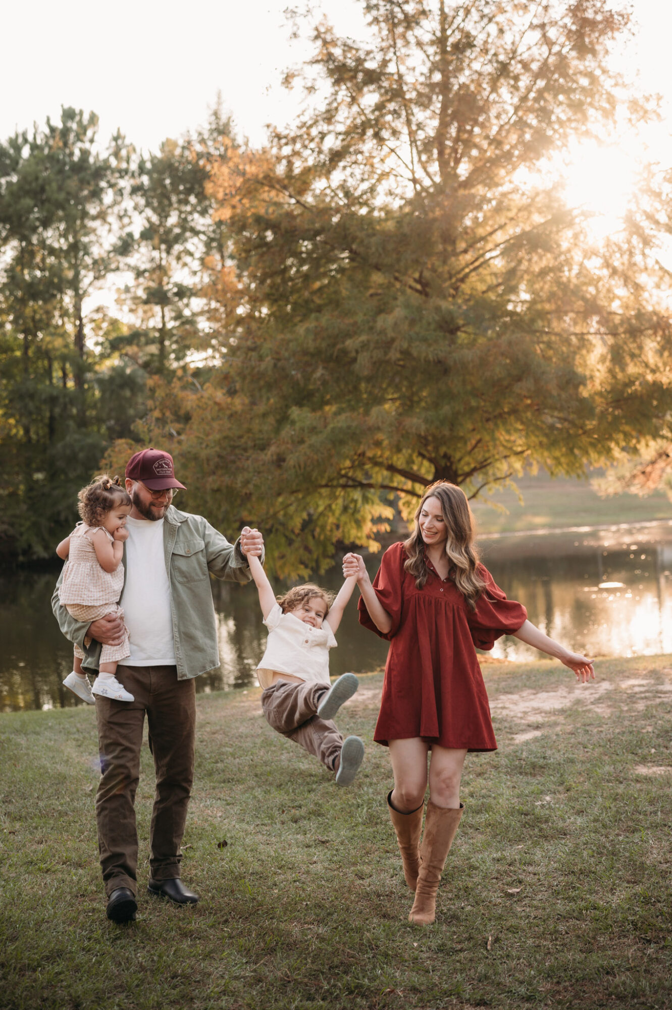 Family of three walking outdoors near a lake with trees, holding hands and playing, sunlight filtering through trees.