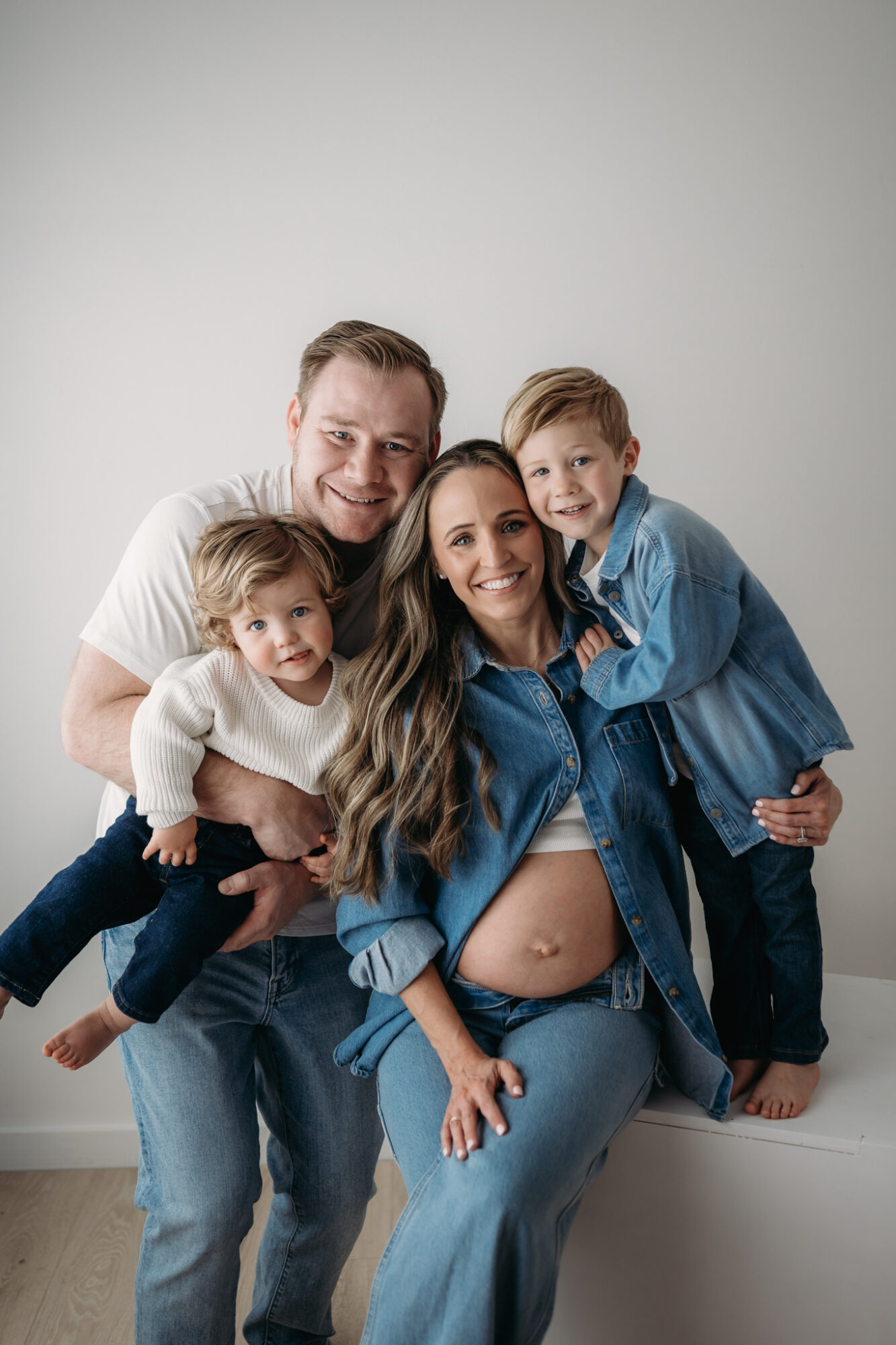Family of four, including pregnant woman, smiling and posing together against a plain wall.