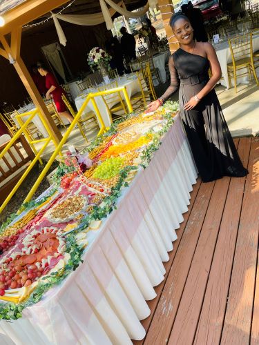 Woman in black dress standing next to a table with colorful food and desserts outdoors.