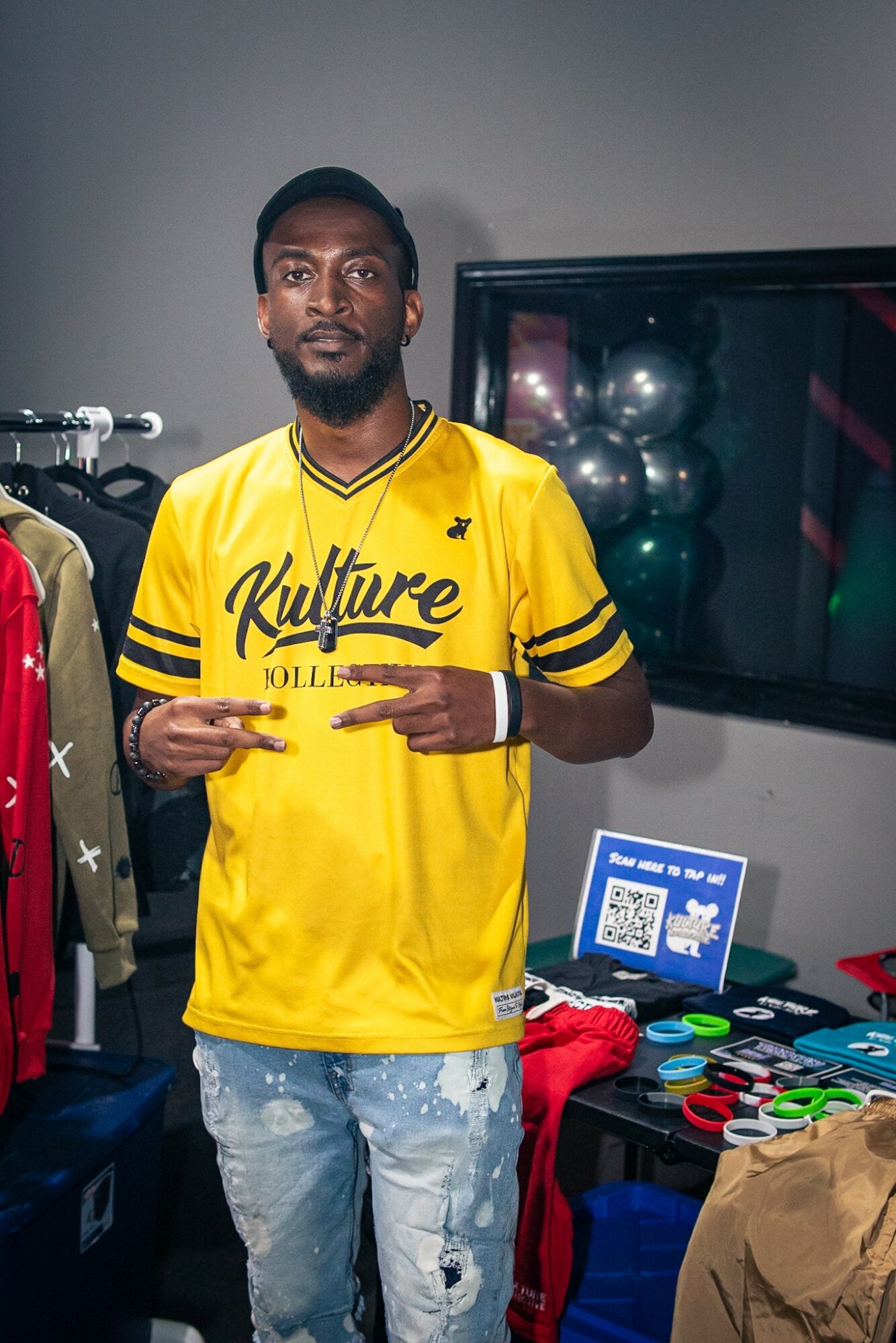 Man wearing a yellow shirt with black text, standing indoors with clothing and a display screen behind him.