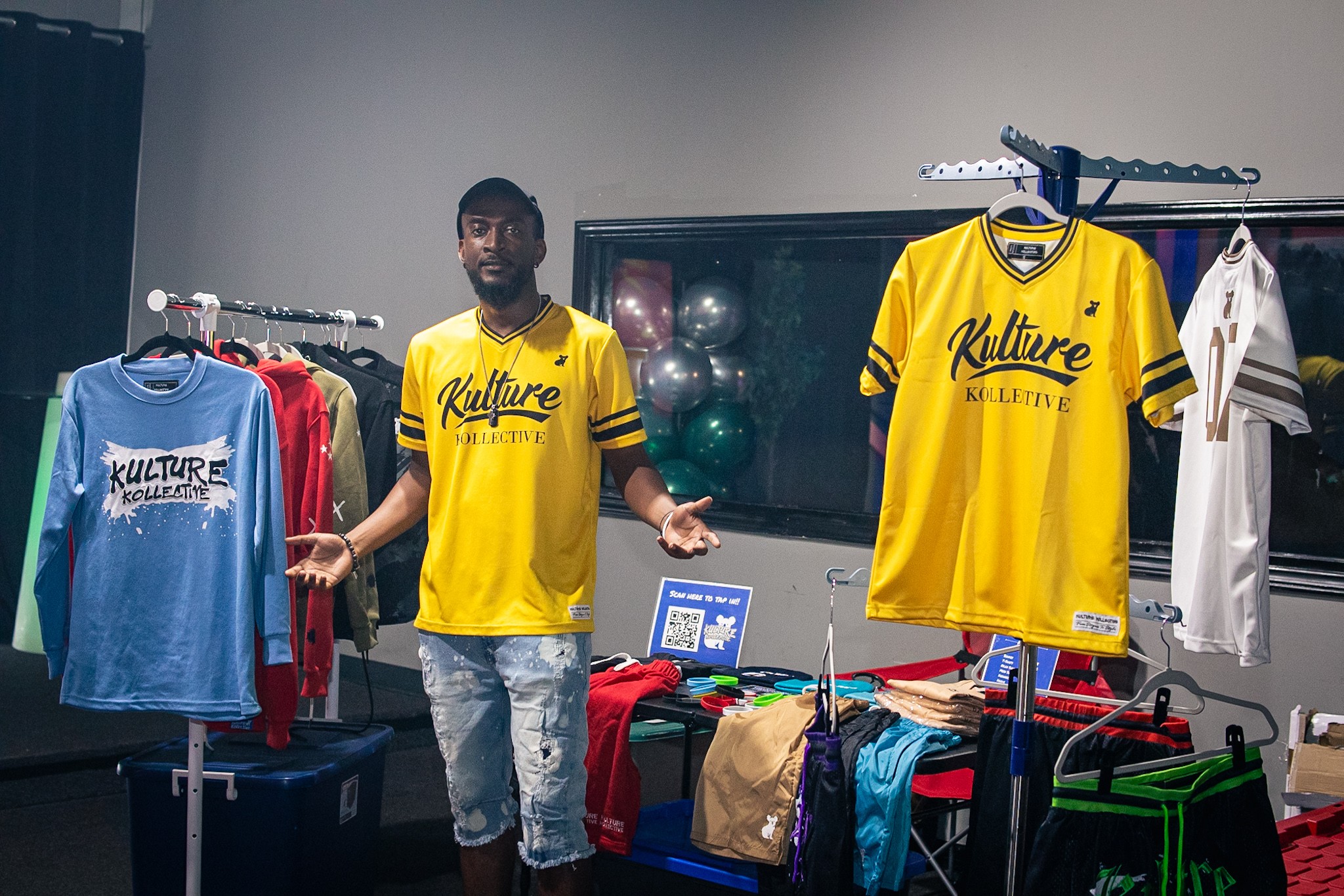 Man standing next to clothing racks with yellow and blue shirts, in a room with sports equipment and tables.