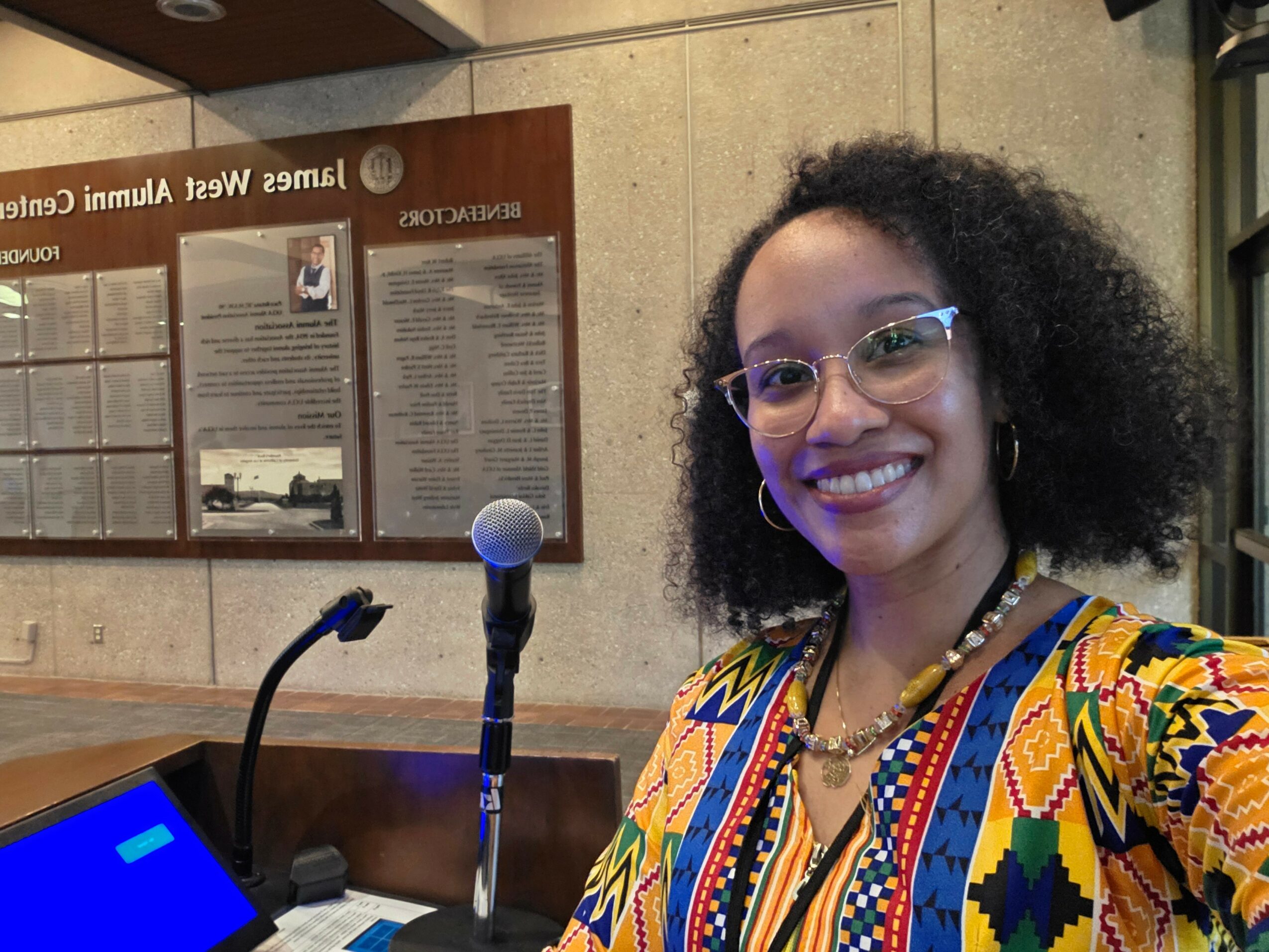 Woman with curly hair and glasses smiling, wearing a colorful patterned top, standing in front of informational boards.