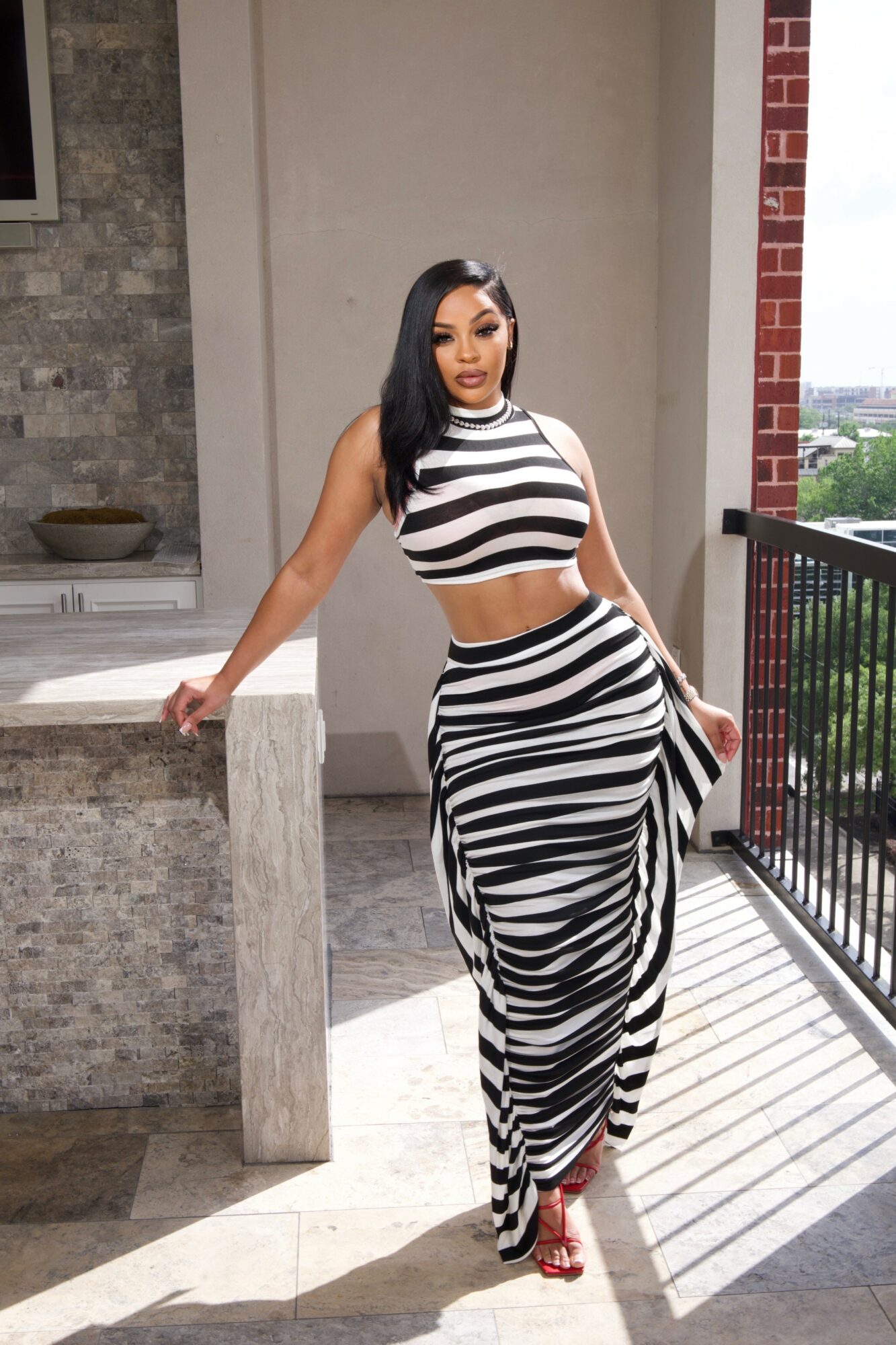 Woman in striped crop top and long skirt standing on balcony with city view, hand on railing, sunlight casting shadows.