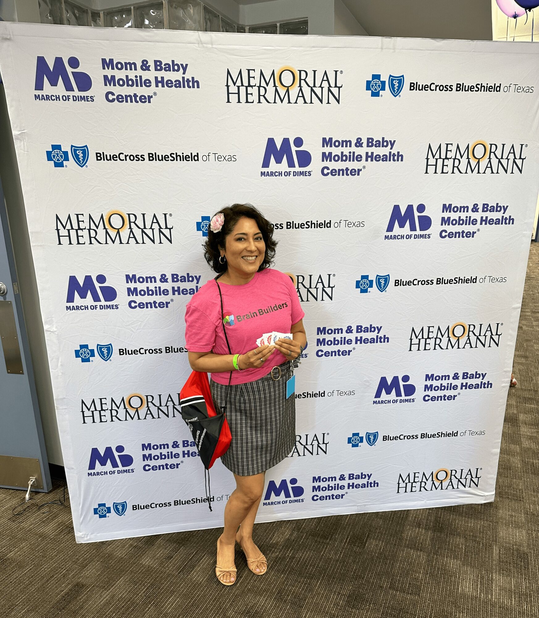 Woman in pink top and striped skirt standing in front of a backdrop with logos and text, smiling, holding a phone.