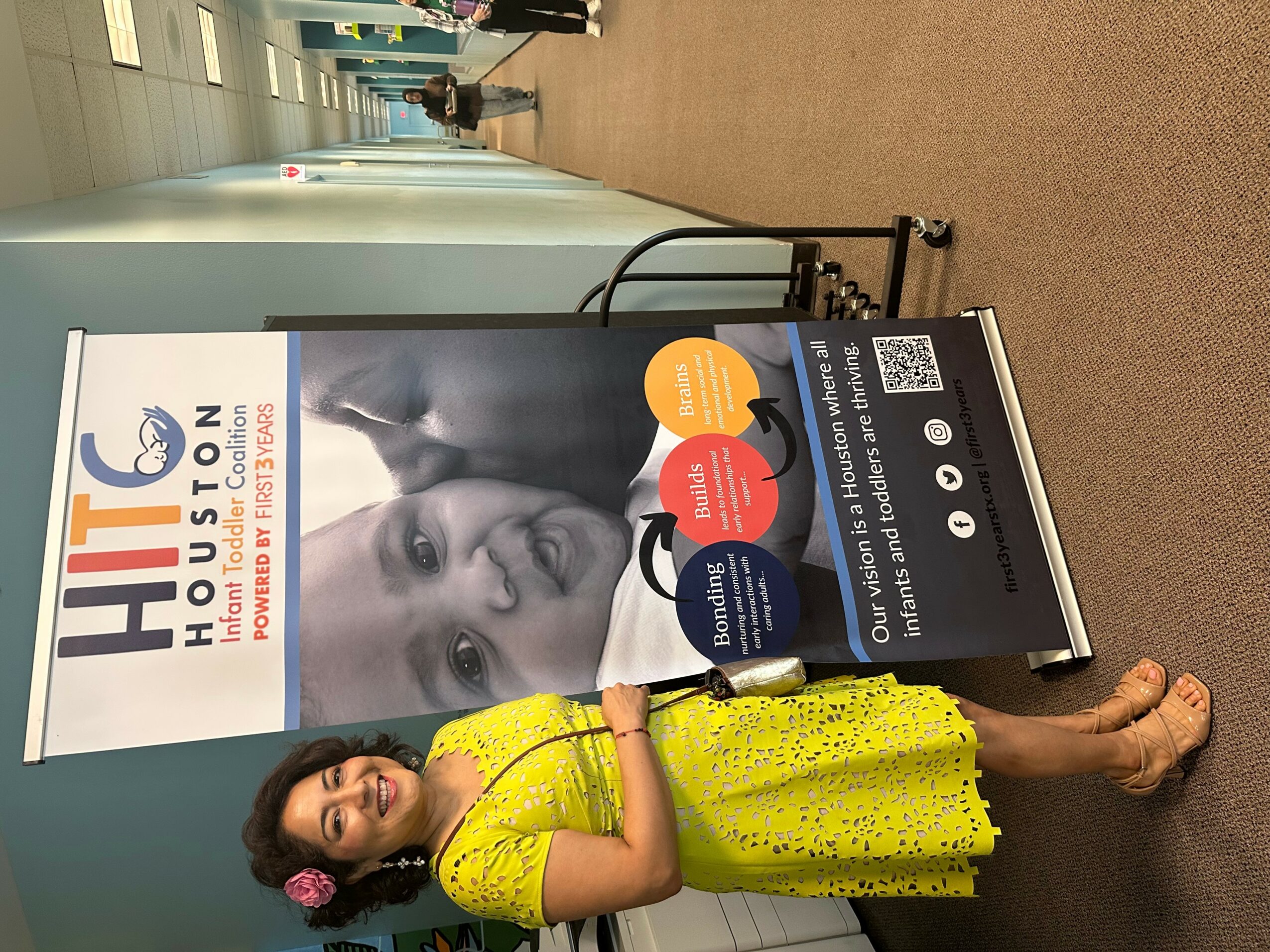 Woman in yellow dress holding a signboard with promotional material, standing in hallway with posters and a carpeted floor.