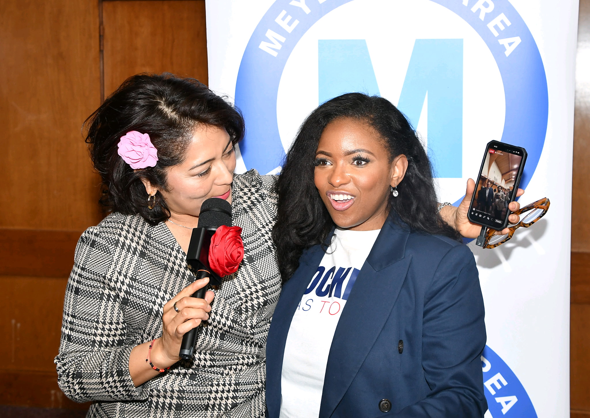 Two women speaking into microphones, one holding a phone, smiling, with a blue and white background featuring a logo.