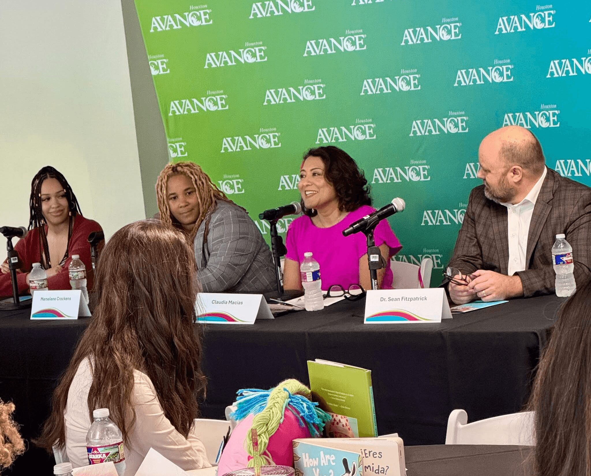 Four people seated at a panel discussion table with microphones and water bottles, in front of a green and blue backdrop.