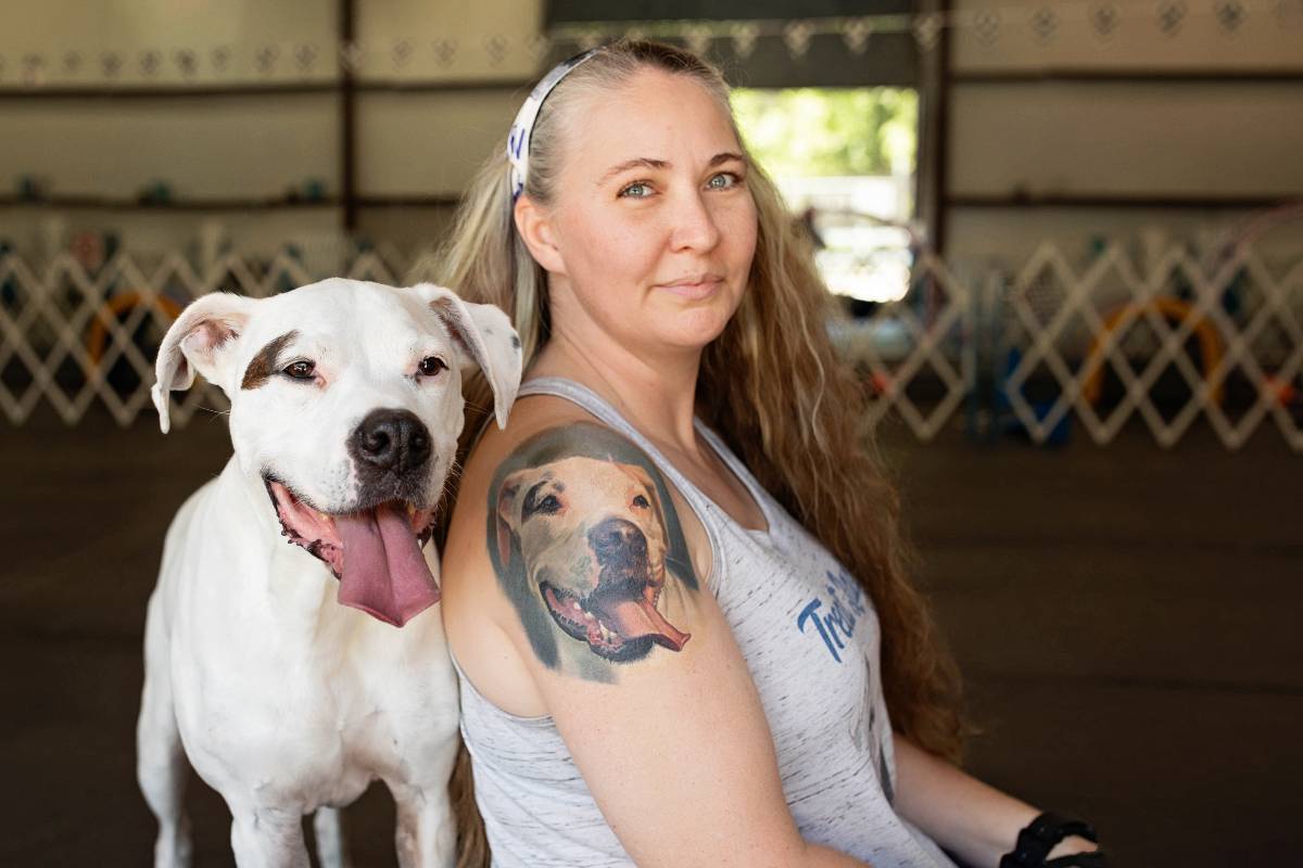Woman with long hair and tattoo of a dog on her arm, smiling next to a white dog with brown spots inside a building.