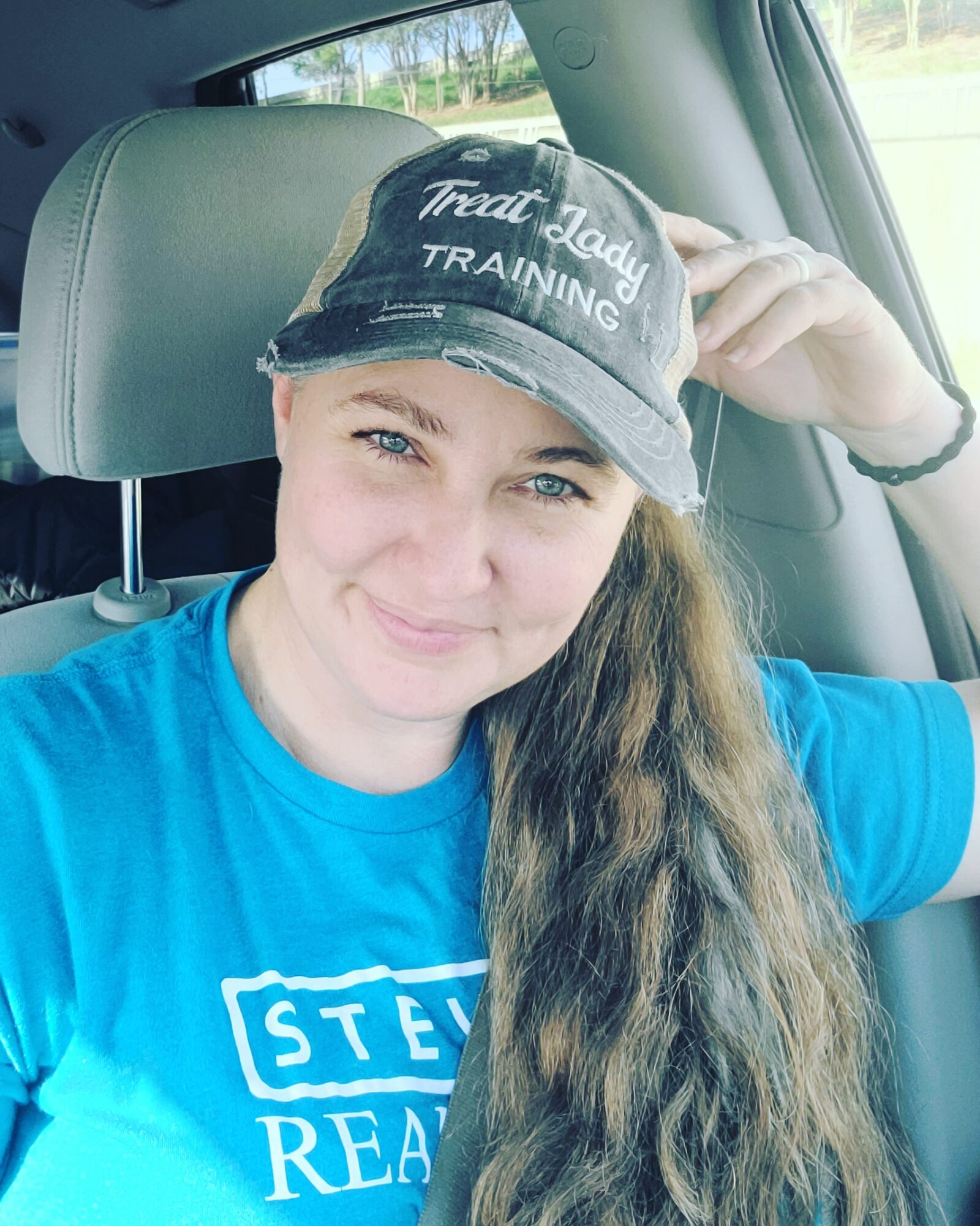 Woman wearing a cap and blue shirt sitting in car, smiling, with long wavy hair.