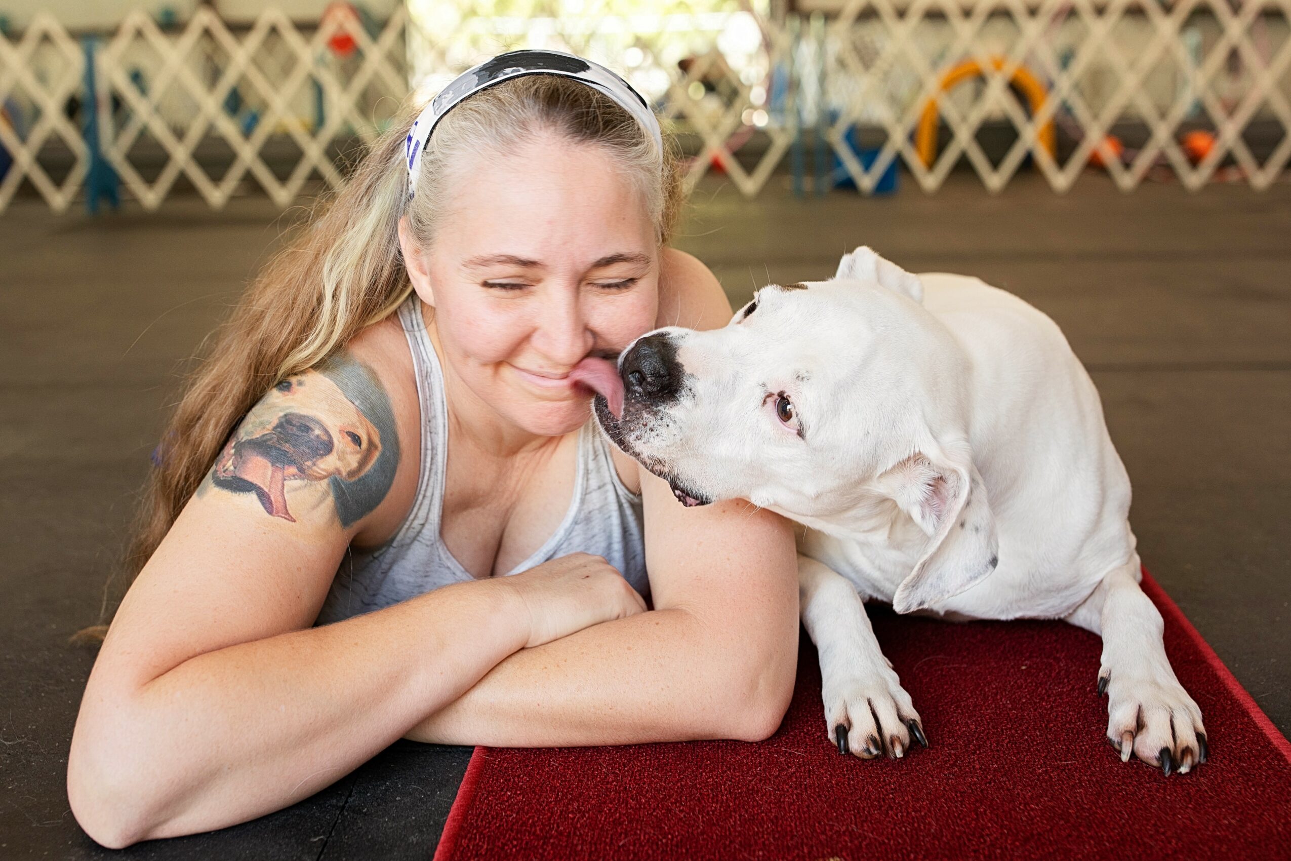 Girl with long hair and a tattoo on her arm smiling with eyes closed, resting her head on her crossed arms, next to a white dog.