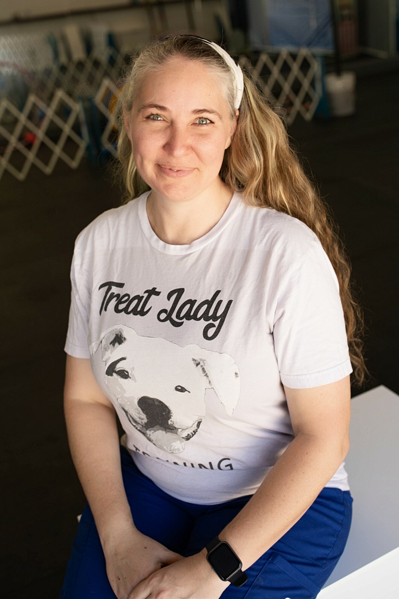 Smiling woman with long wavy hair, wearing a white T-shirt and blue pants, sitting indoors.