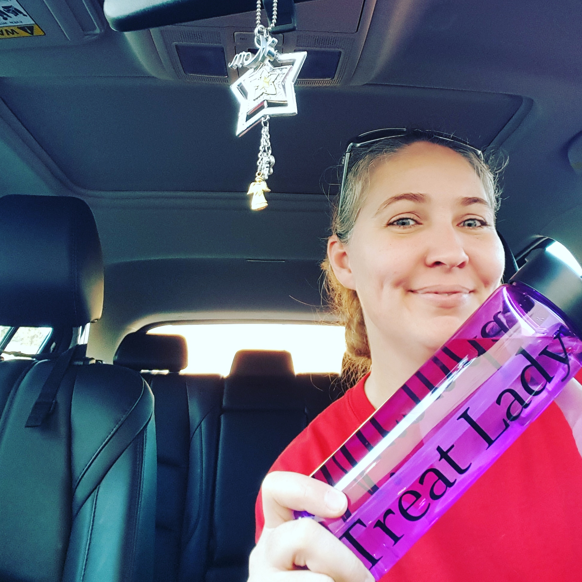 Young woman with glasses on head smiling inside a car, holding a pink water bottle with 'Treat Lady' written on it.