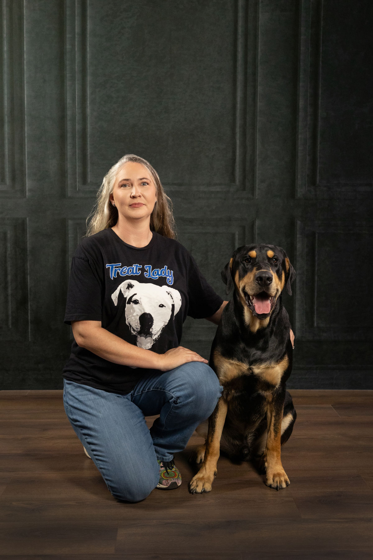 Woman kneeling next to a large Rottweiler dog indoors, dark wall background, woman wearing a black T-shirt with a bear graphic.