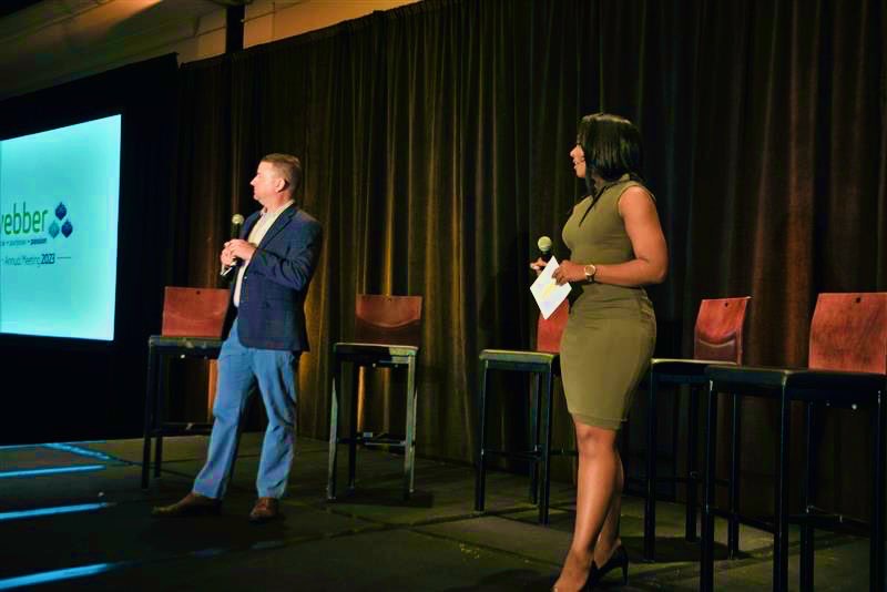 Two people on stage, one speaking into a microphone, the other holding papers, with chairs and a screen behind them.