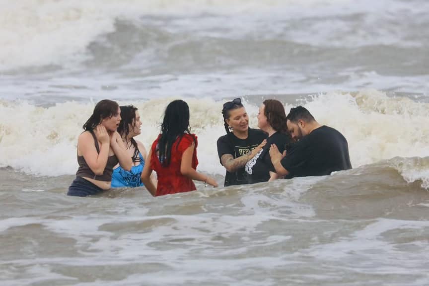 Group of six people standing in ocean waves, engaging in conversation and smiling.