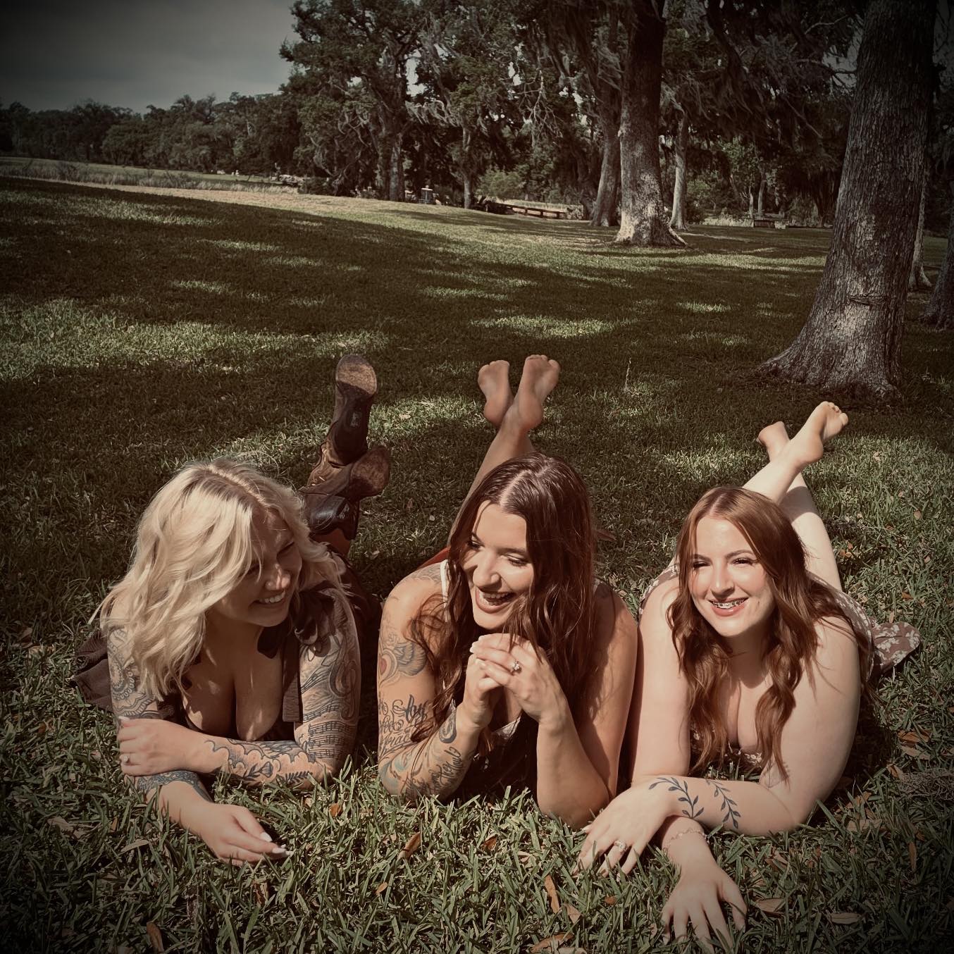 Three women lying on grass with trees in background, smiling and laughing together outdoors.