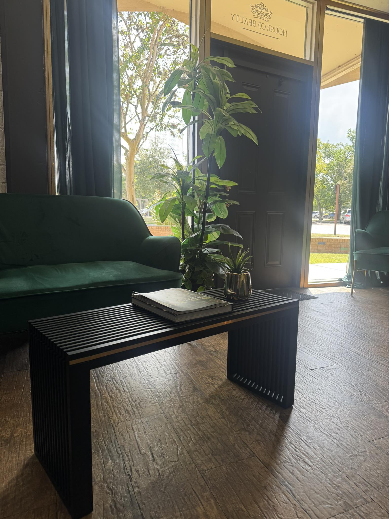 Indoor space with a dark wooden table, green sofa, tall plant, and large window showing outdoor trees and sunlight.
