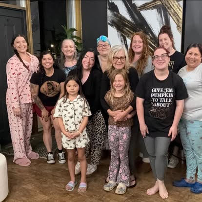 Group of fifteen women and children standing indoors, smiling, in front of abstract artwork and plants.