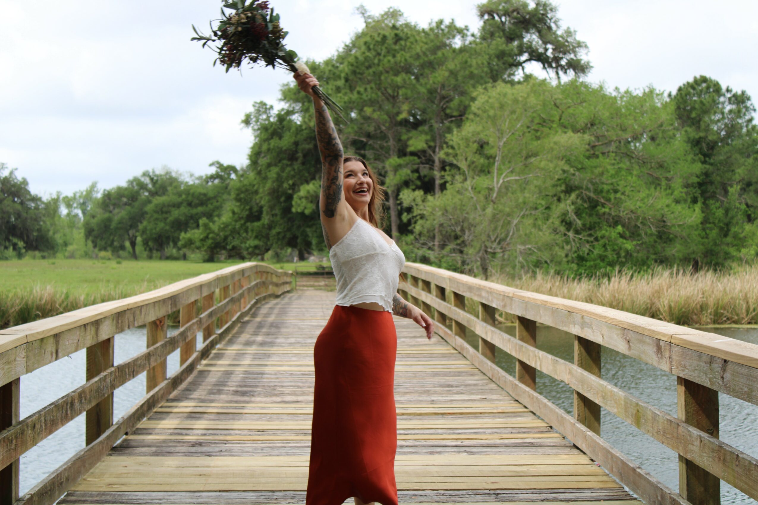 Woman standing on a wooden bridge holding a bouquet of flowers above her head, smiling, with trees in the background.