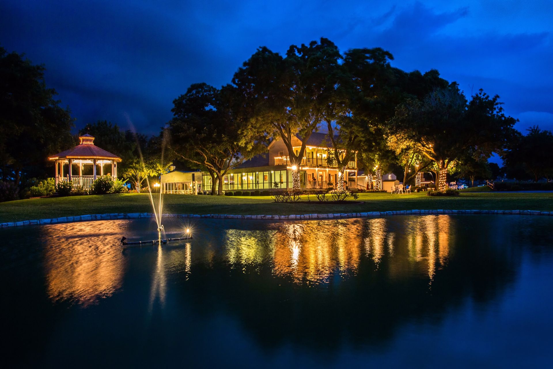 Night scene of a park with a pond, illuminated trees, and a lit building in the background.