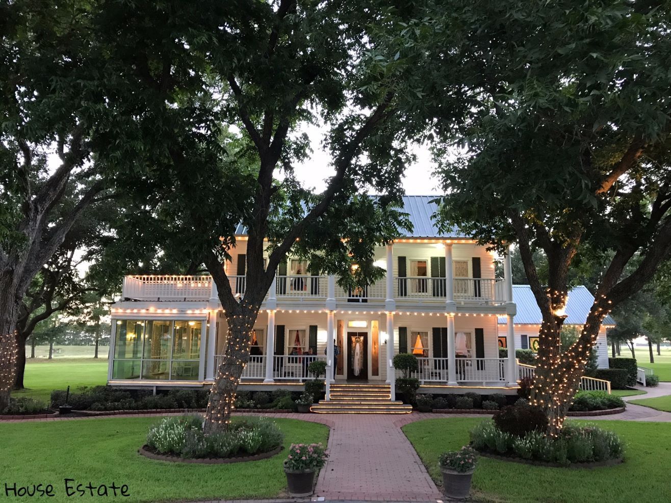 Large house with multiple balconies, surrounded by trees with string lights, front walkway, and landscaped yard.