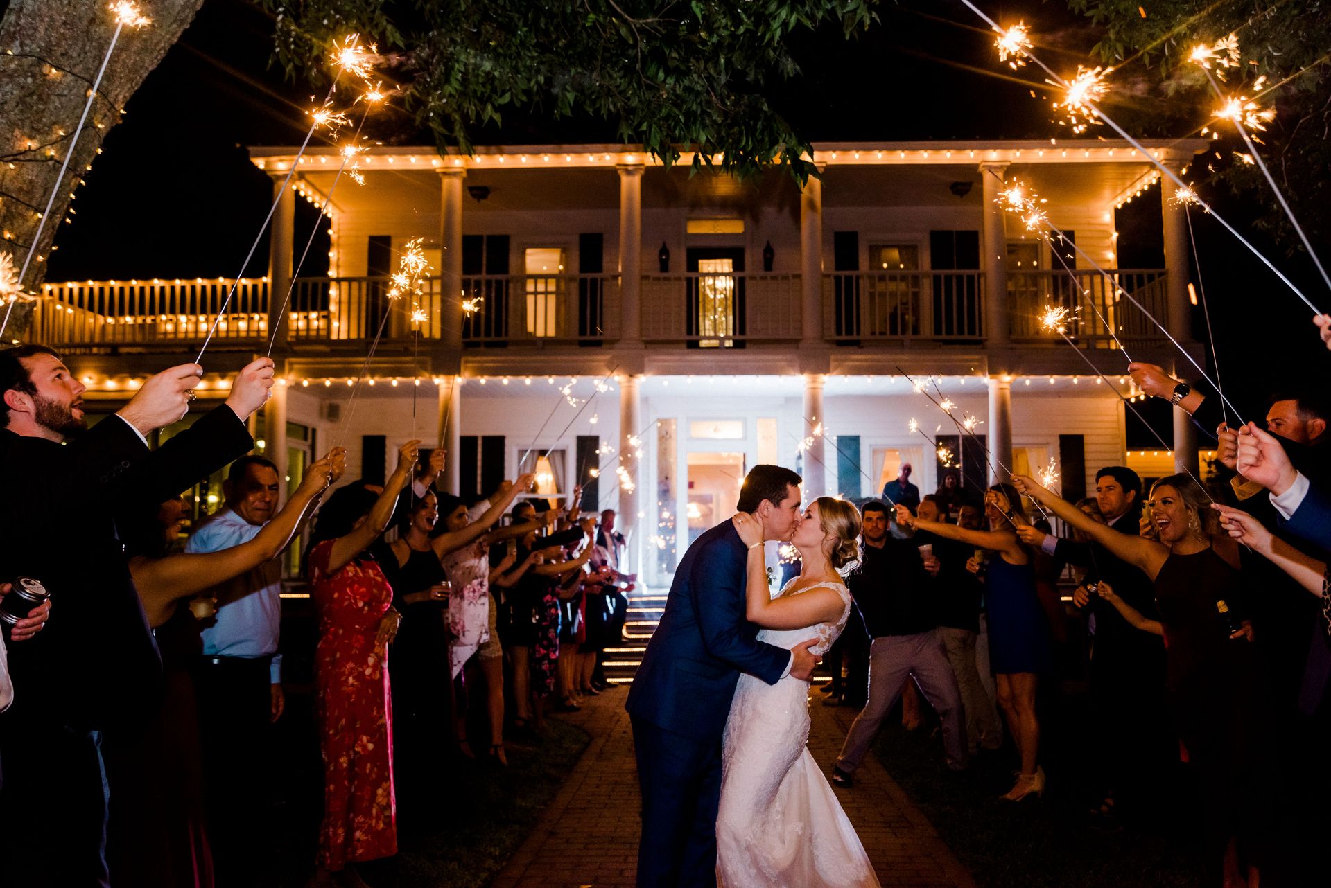 Couple kissing in front of a lit building with guests holding sparklers during night celebration.