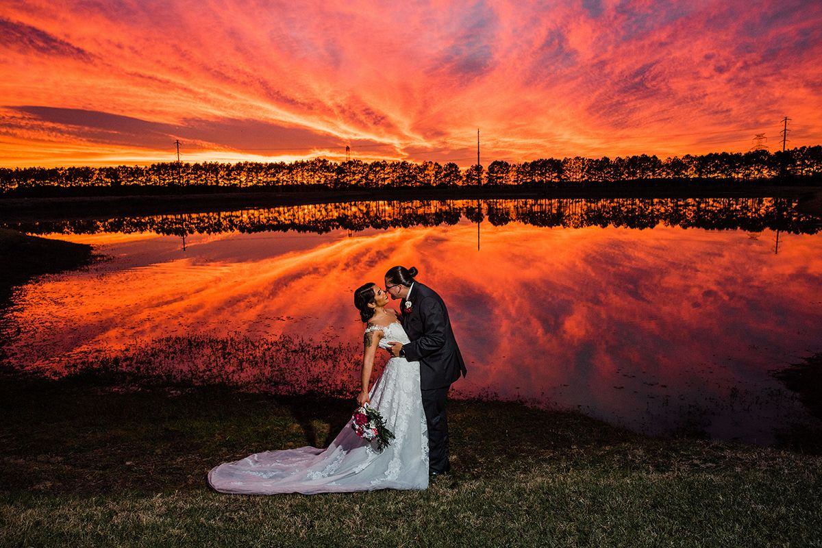 Couple in wedding attire standing by a lake during sunset with vibrant orange and pink sky reflected in water.
