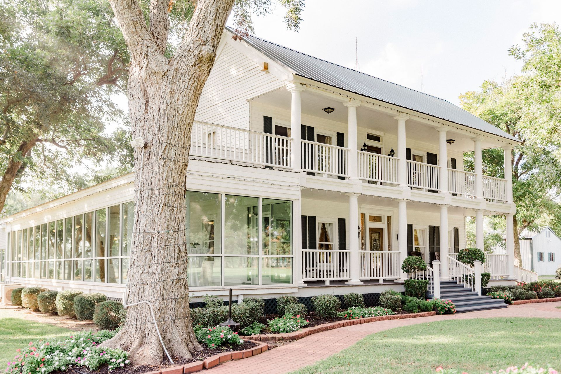 Two-story house with a large porch, surrounded by trees and landscaped yard, with a glass-enclosed extension on the side.